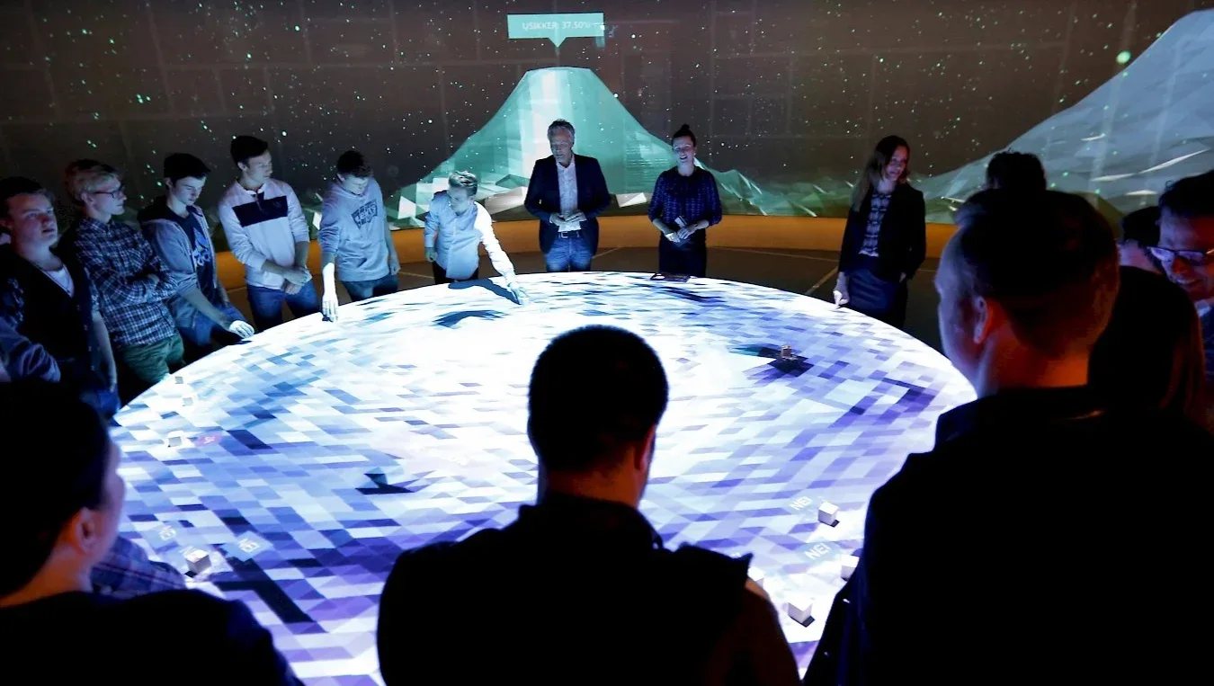 Group of people gathered around interactive digital display showing blue data visualization with mountain imagery backdrop