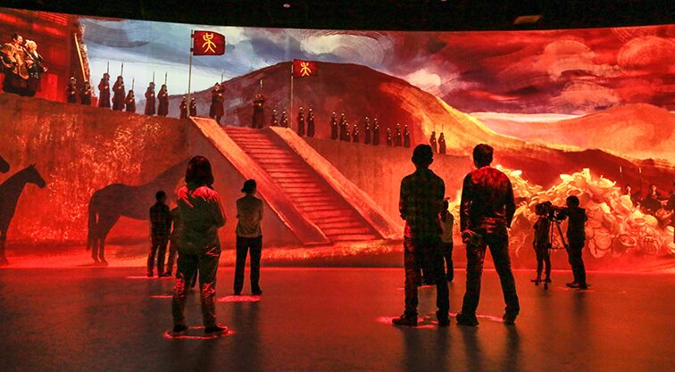 Immersive red-lit digital installation depicting silhouetted figures viewing massive projected historical scenes.