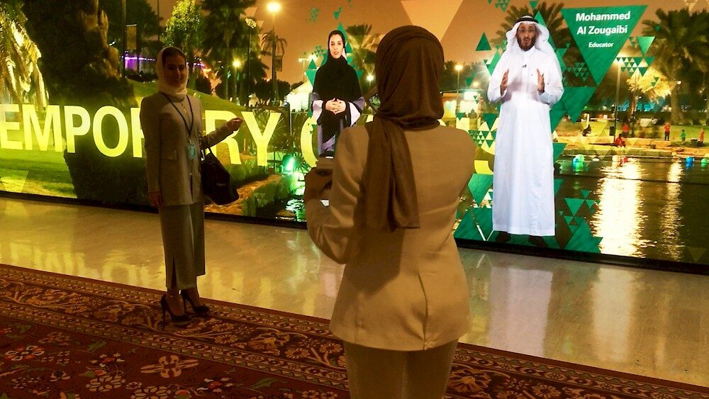 People gathered at illuminated event display featuring educator Mohammed Al Zougaibi in traditional white attire.