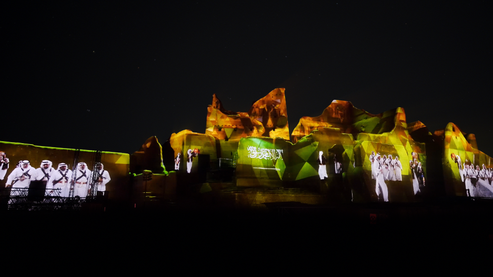 Historic building illuminated at night with projection mapping displaying people and Arabic text.