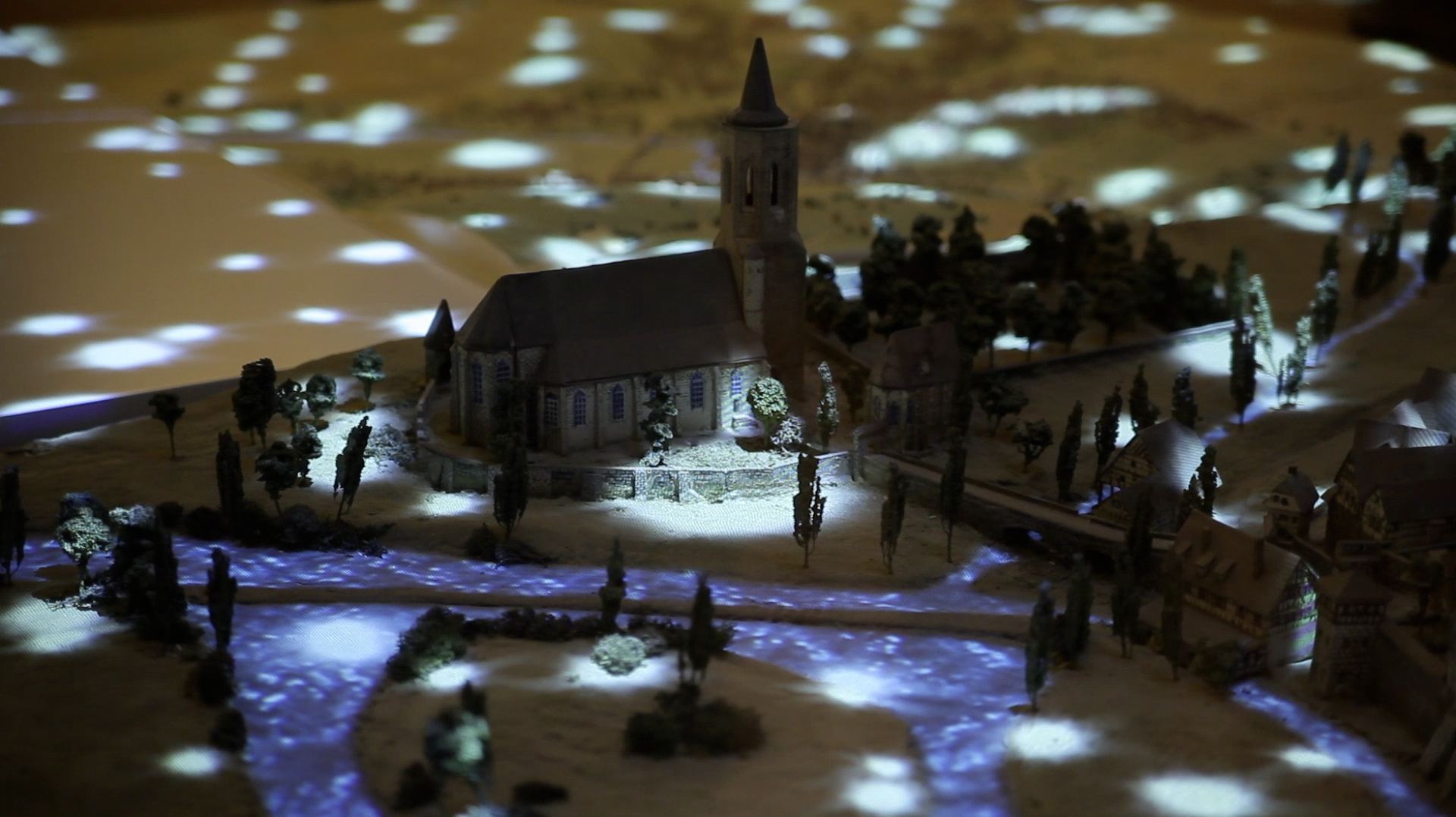 Miniature snow-covered village with illuminated church, cypress trees, and glowing blue water at night