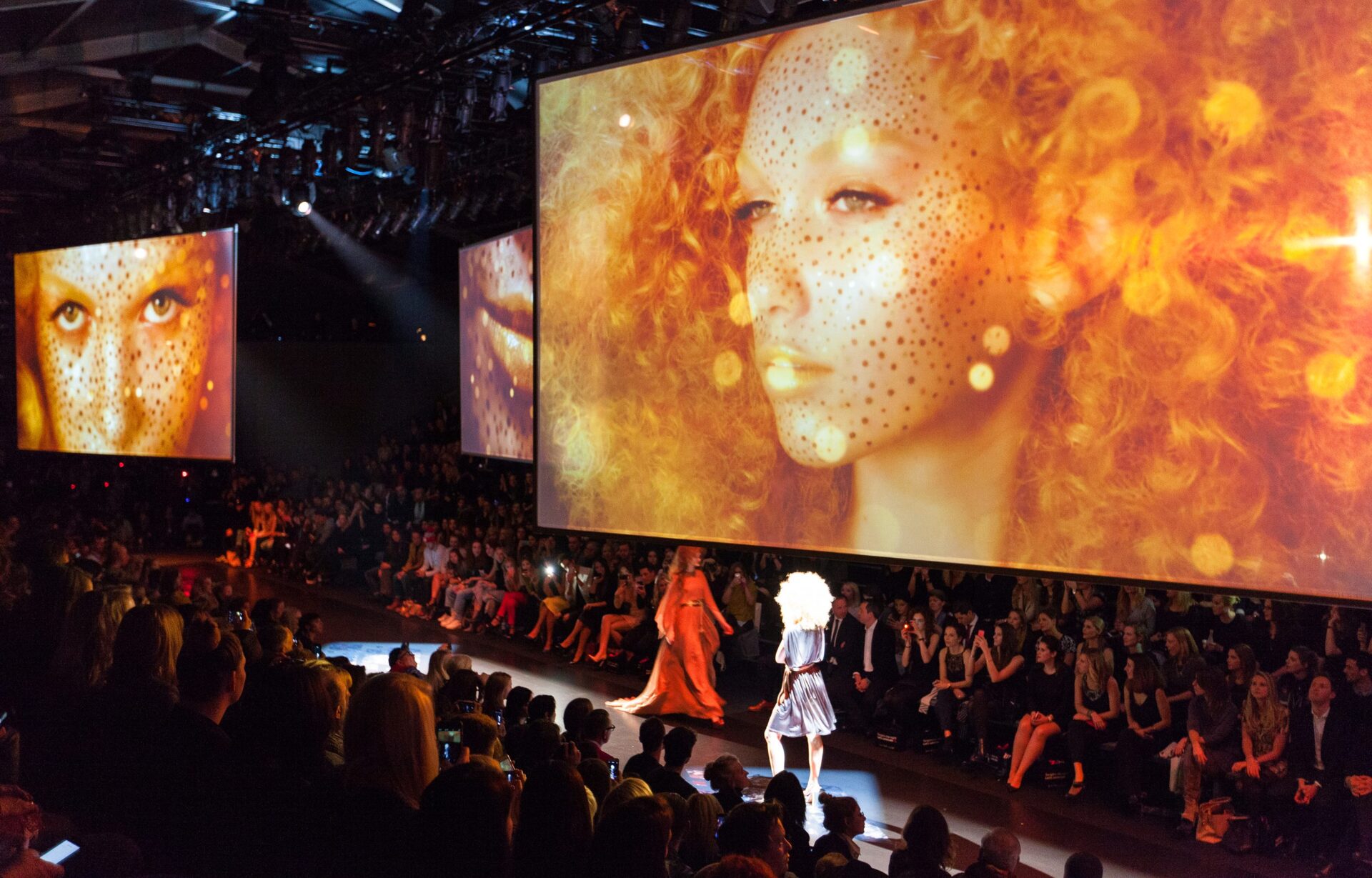 Fashion show runway with models and large orange-toned portrait displays on screens