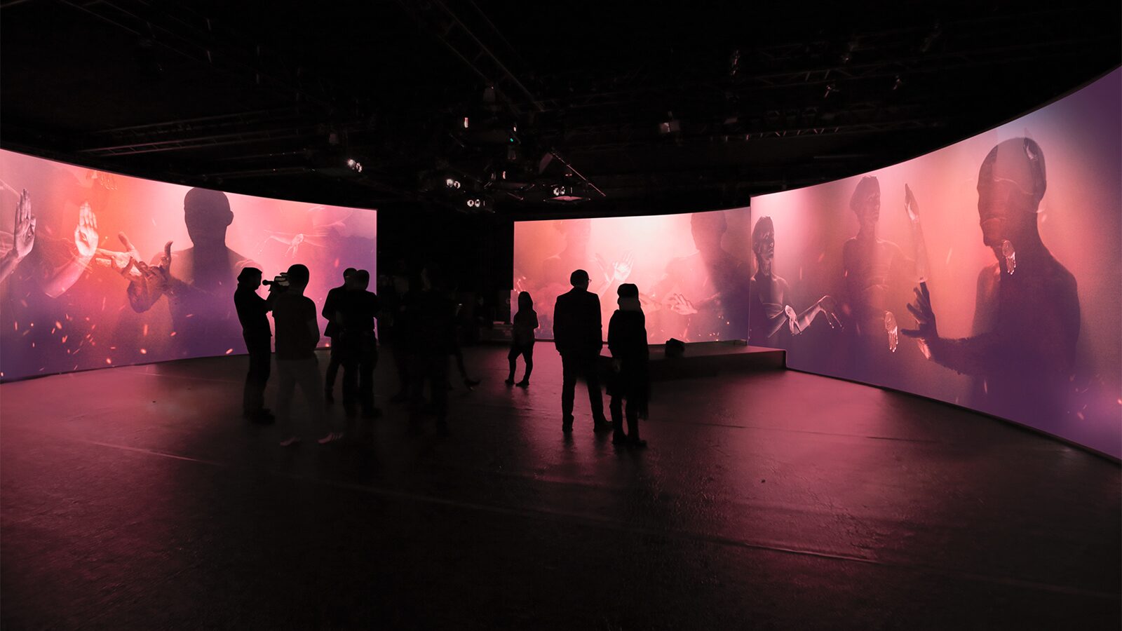 Silhouetted visitors observe immersive pink video installation with life-size projected figures.