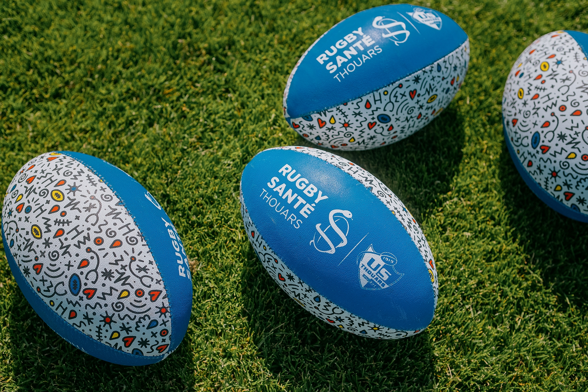 Quatre ballons de rugby bleus avec motifs colorés posés sur l'herbe, marqués Rugby Santé Thouars.