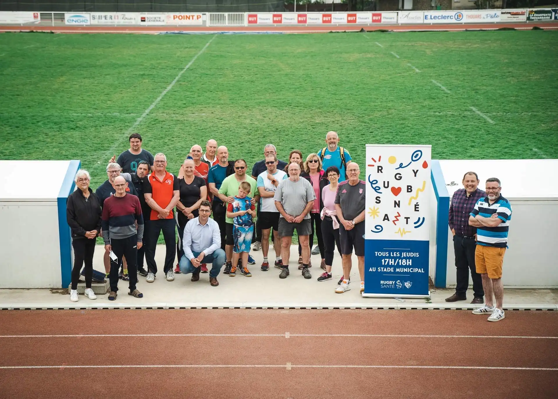 Un groupe de personnes de tous âges posant sur une piste d'athlétisme devant un panneau signalant une activité de rugby-santé 