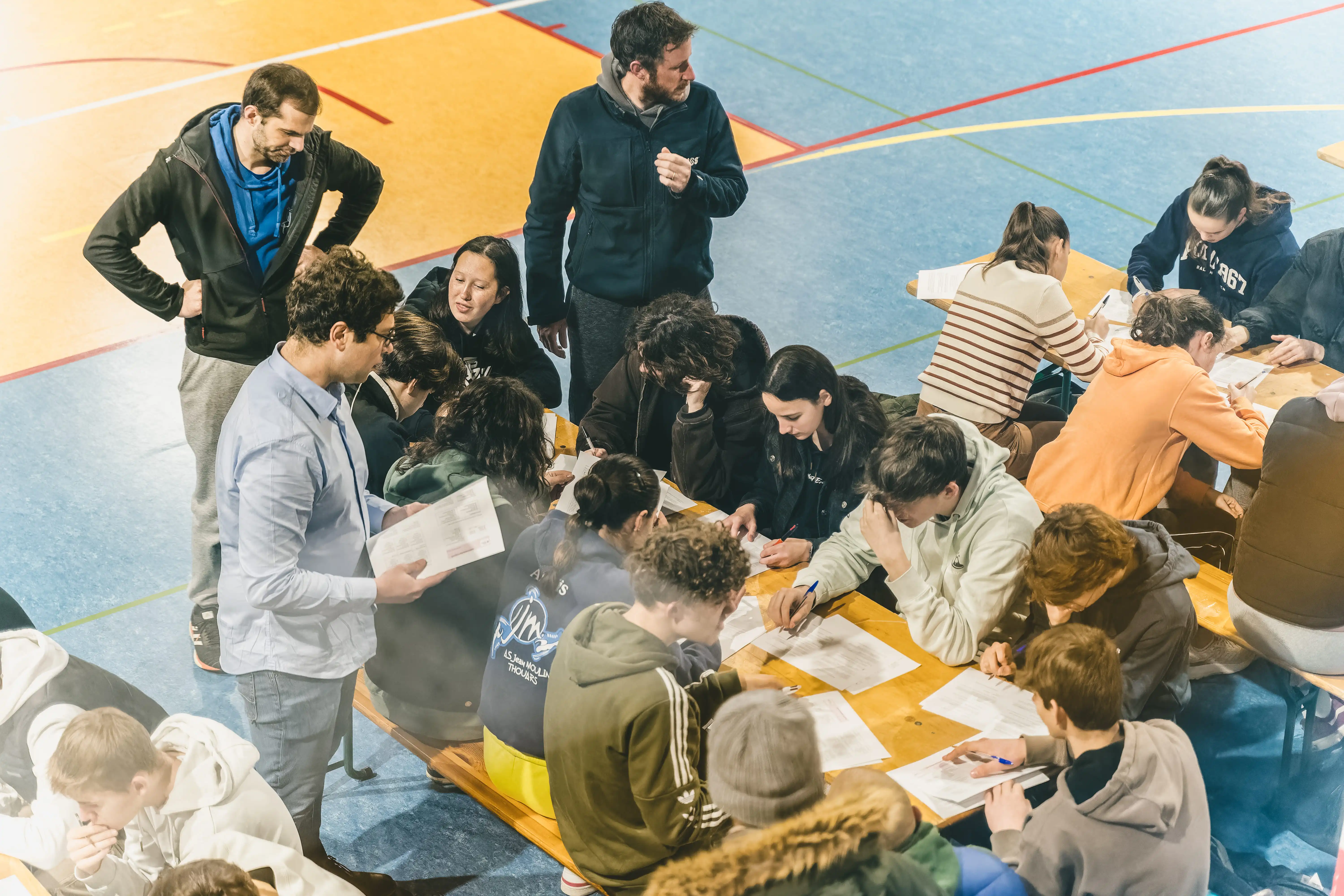Groupe d'étudiants assis à des tables dans un gymnase en train de remplir des formulaires, encadrés par deux adultes debout.