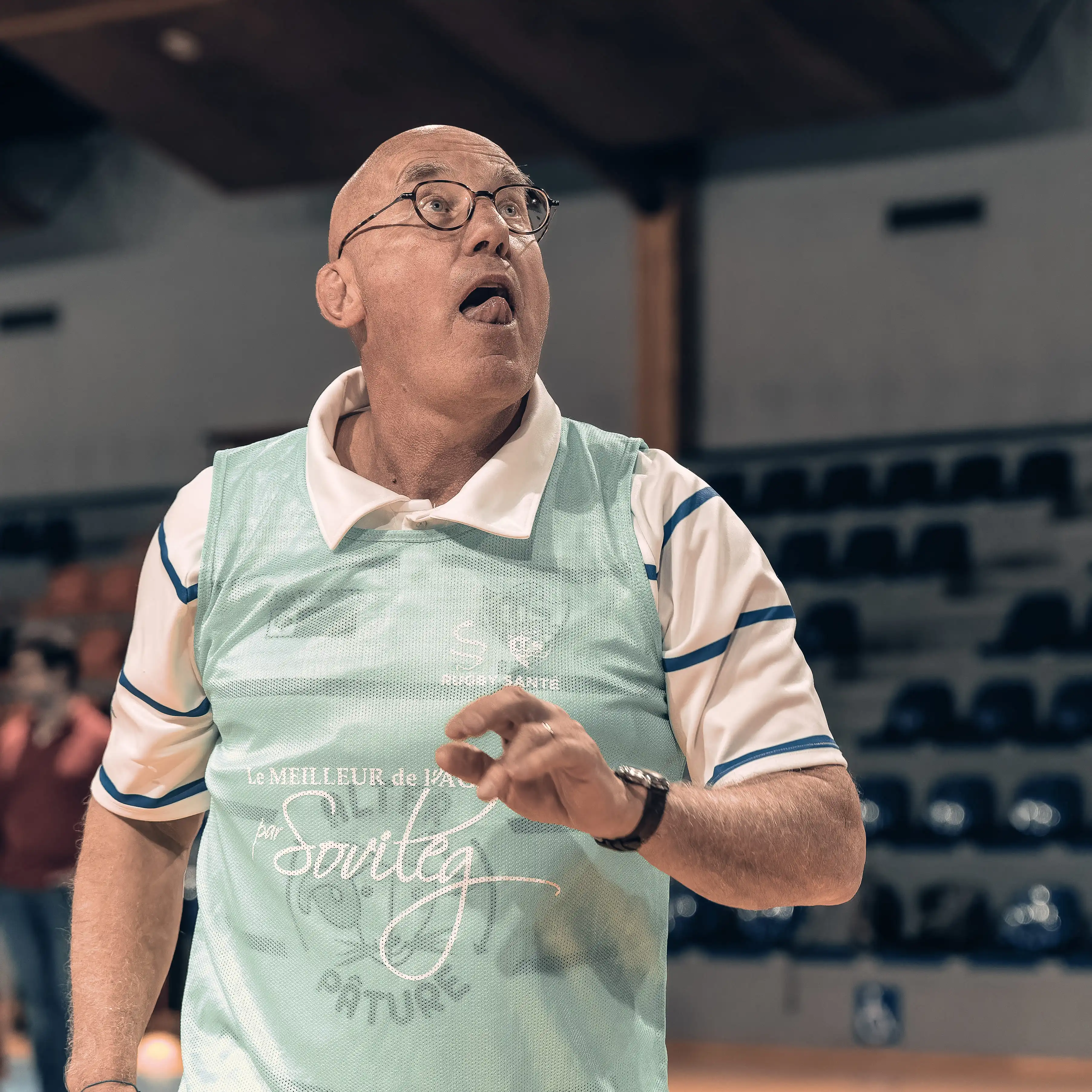 Un homme âgé portant des lunettes et un maillot de sport bleu clair fait un geste de la main avec la bouche ouverte dans un gymnase.