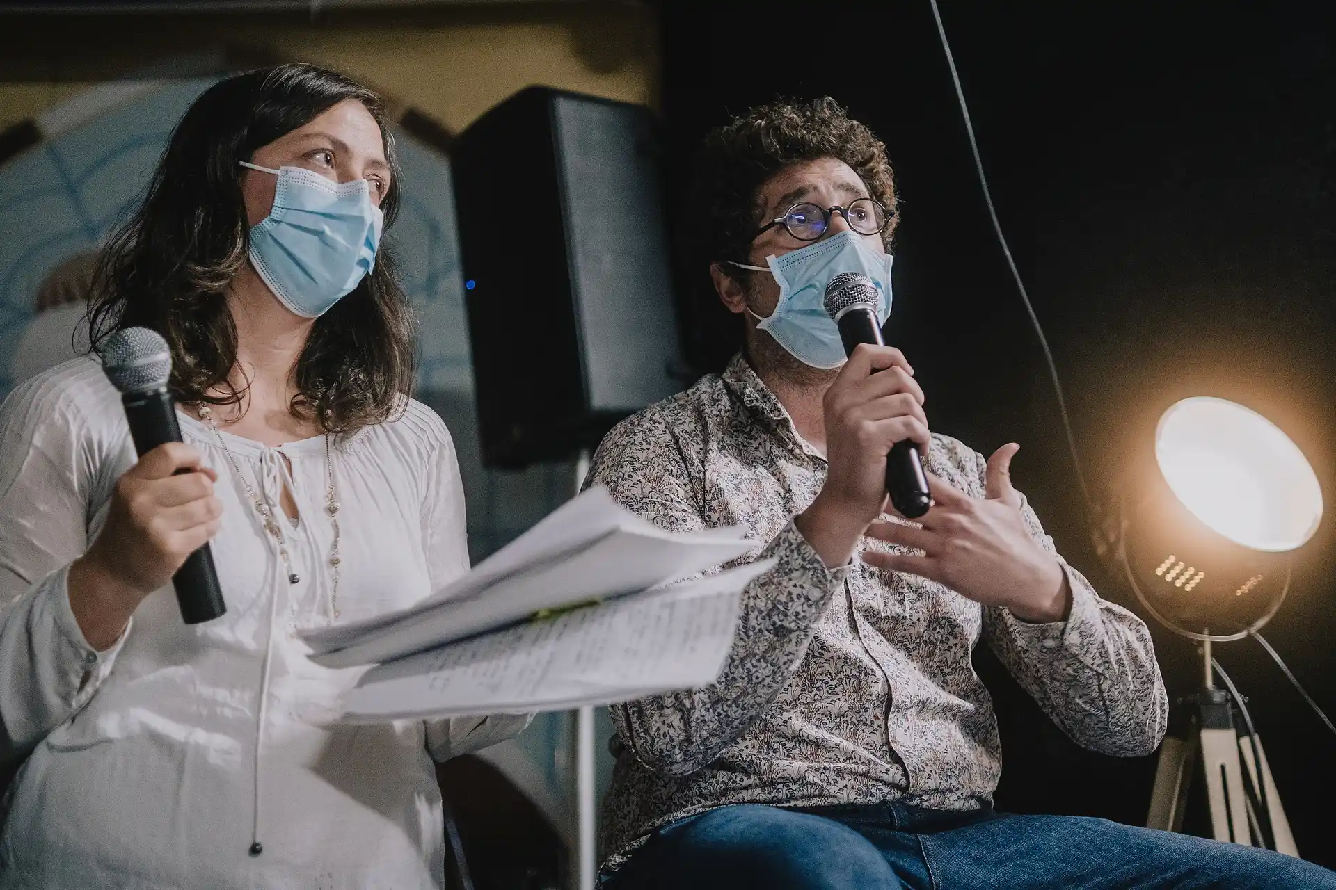 Deux personnes masquées tenant des micros, un homme parlant avec un geste de la main et une femme tenant des feuilles.