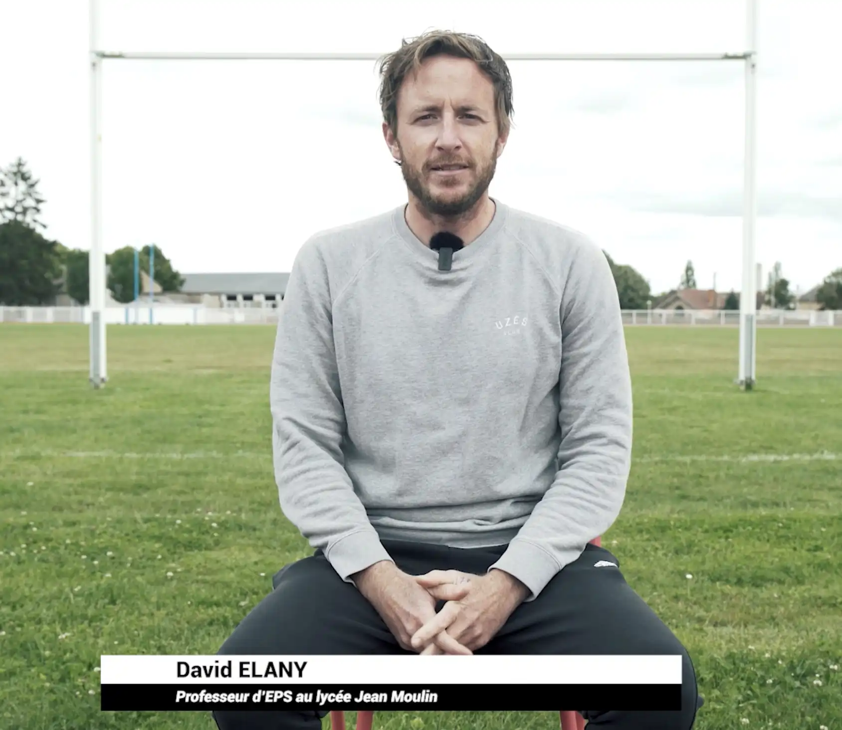 Homme avec un micro-cravate assis sur un terrain de sport, portant un sweat gris et un pantalon noir.