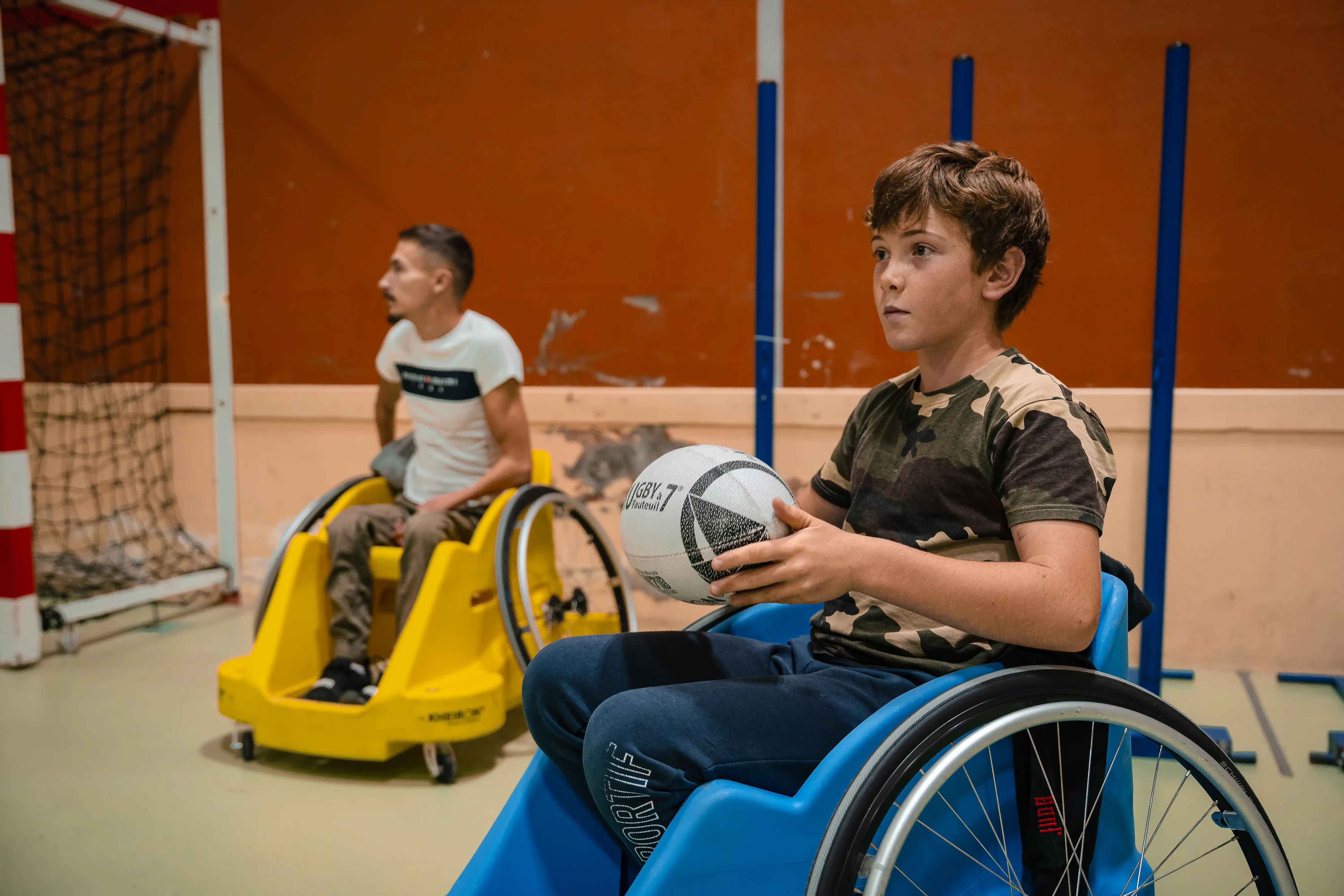 Deux garçons en fauteuils roulants jouent au rugby en salle, l'un tient un ballon de rugby.