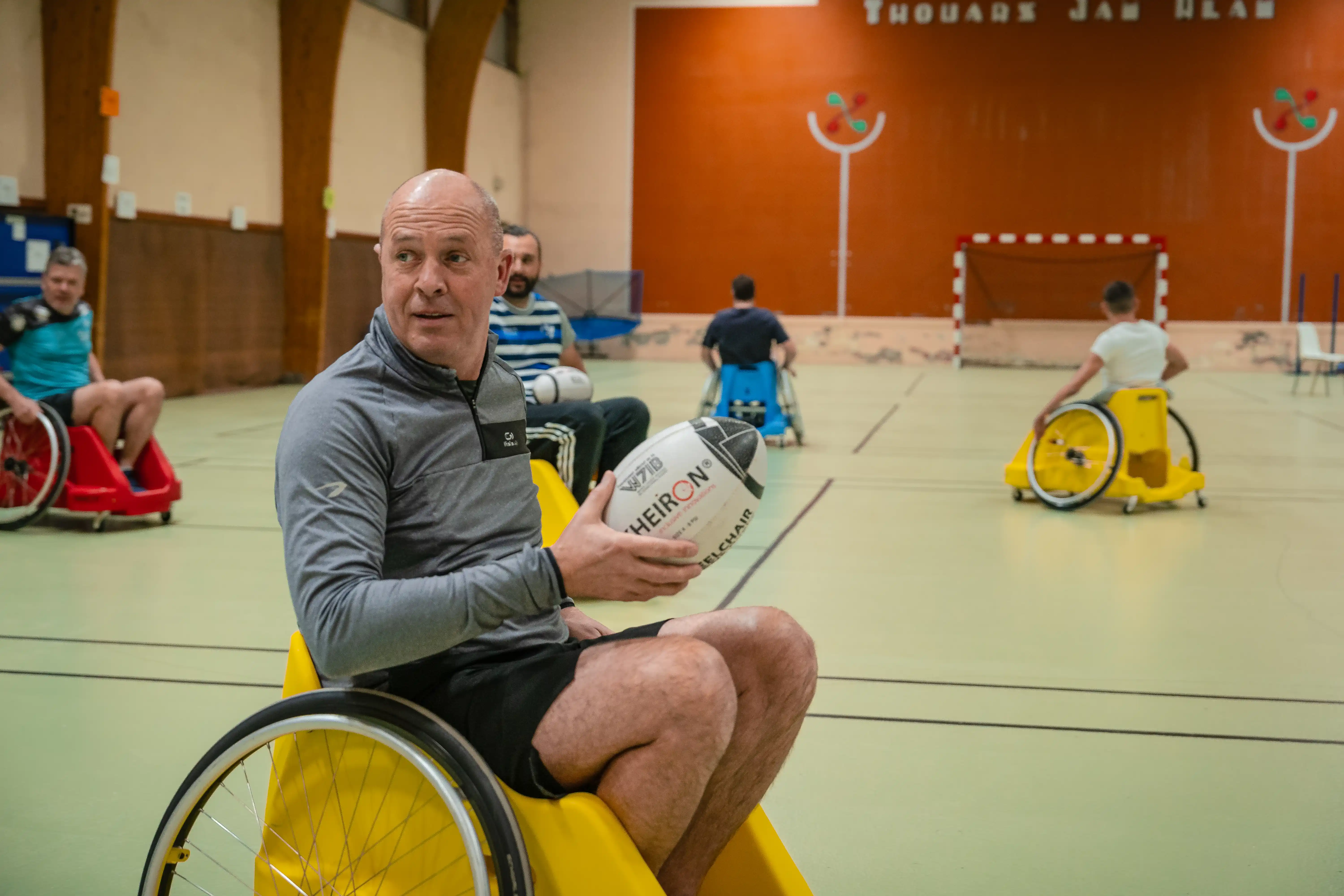 Un homme en fauteuil roulant jaune tenant un ballon de rugby dans un gymnase avec d'autres joueurs en fauteuil roulant jaune et rouge jouant au rugby.