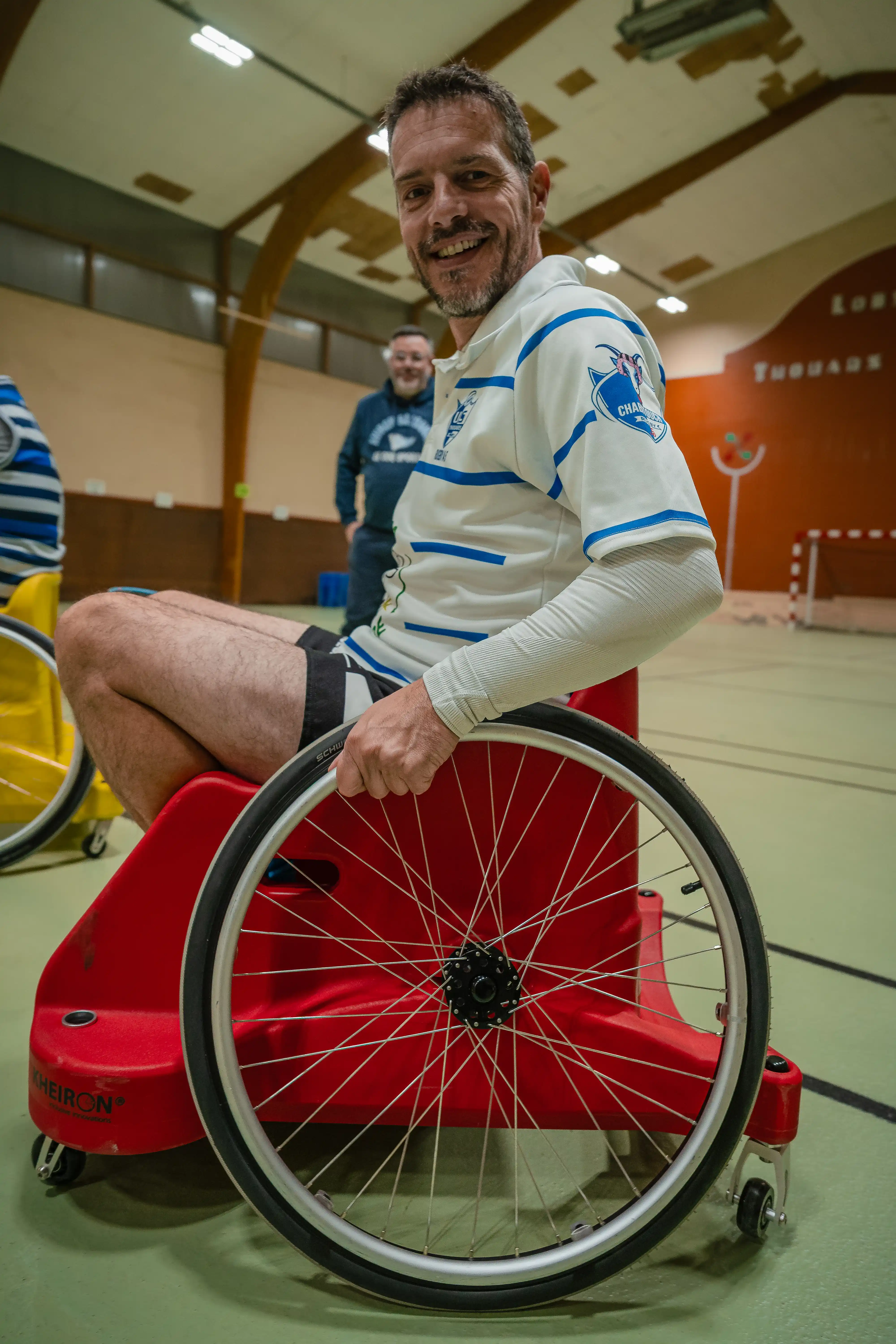 Homme souriant en fauteuil roulant de sport rouge dans une salle de sport intérieure.