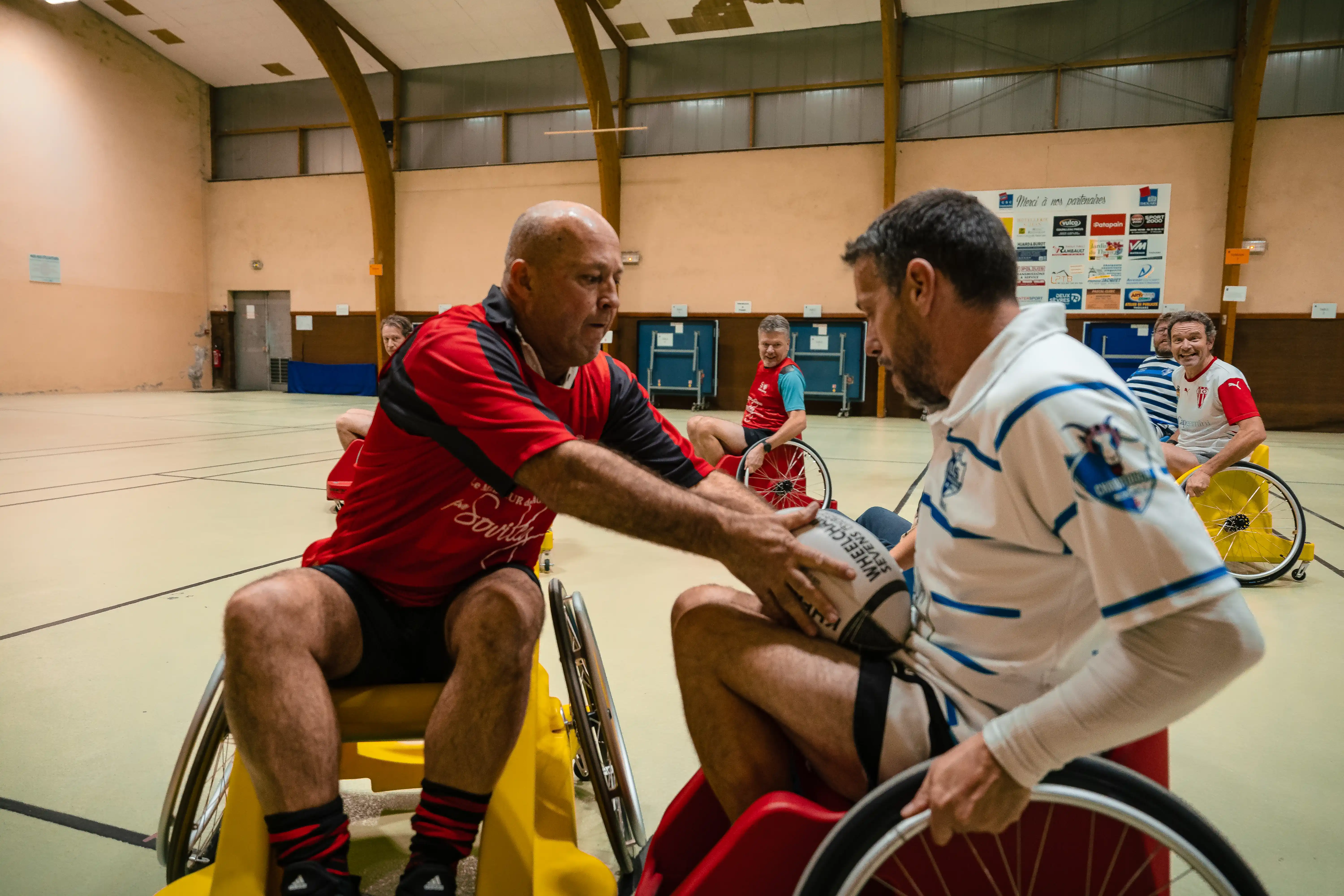 Deux hommes en fauteuil roulant jouent au rugby en intérieur, l'un en maillot rouge passe un ballon à l'autre en maillot blanc.