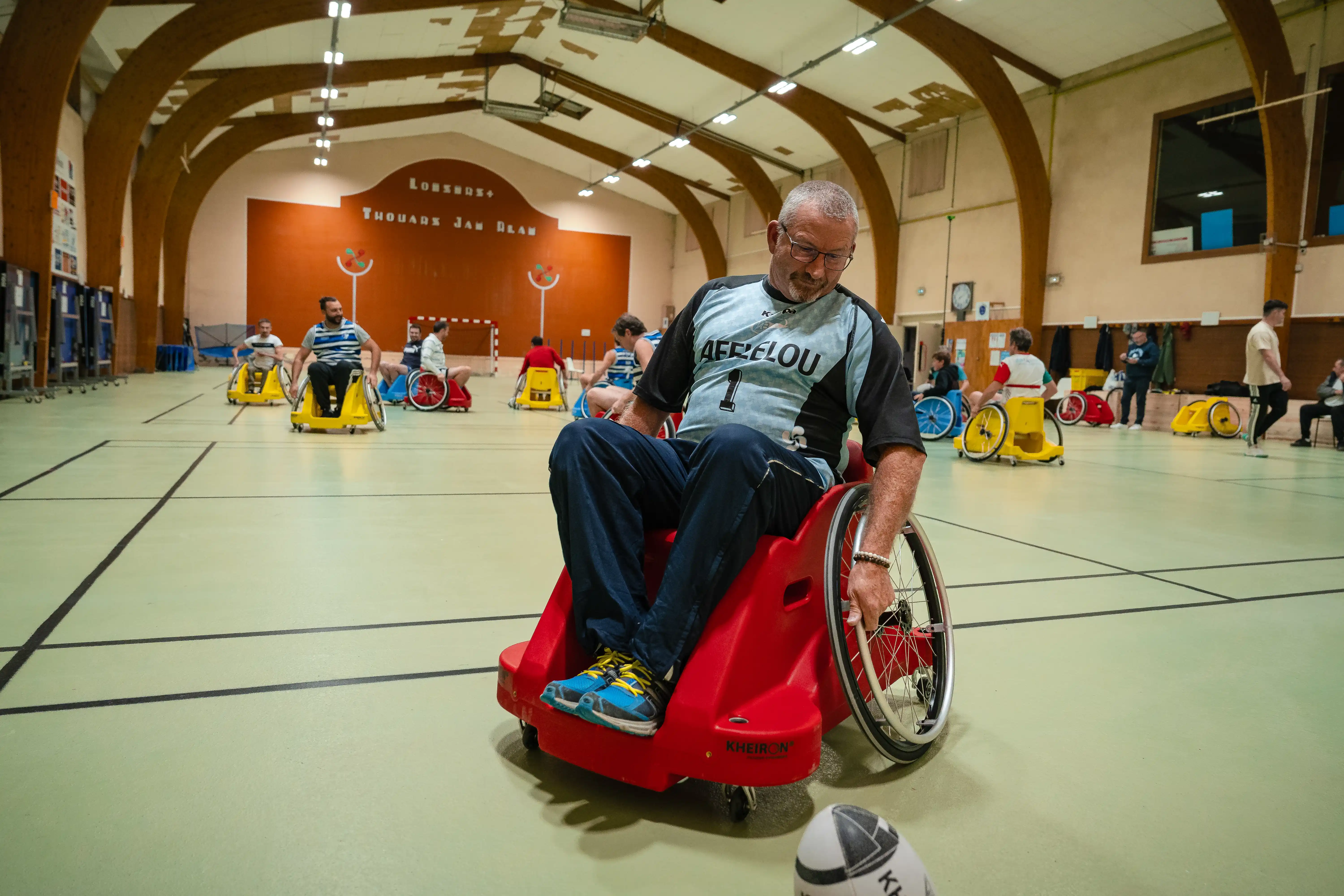 Un homme en fauteuil roulant rouge joue au rugby fauteuil dans une salle de sport avec plusieurs autres joueurs en fauteuils colorés.