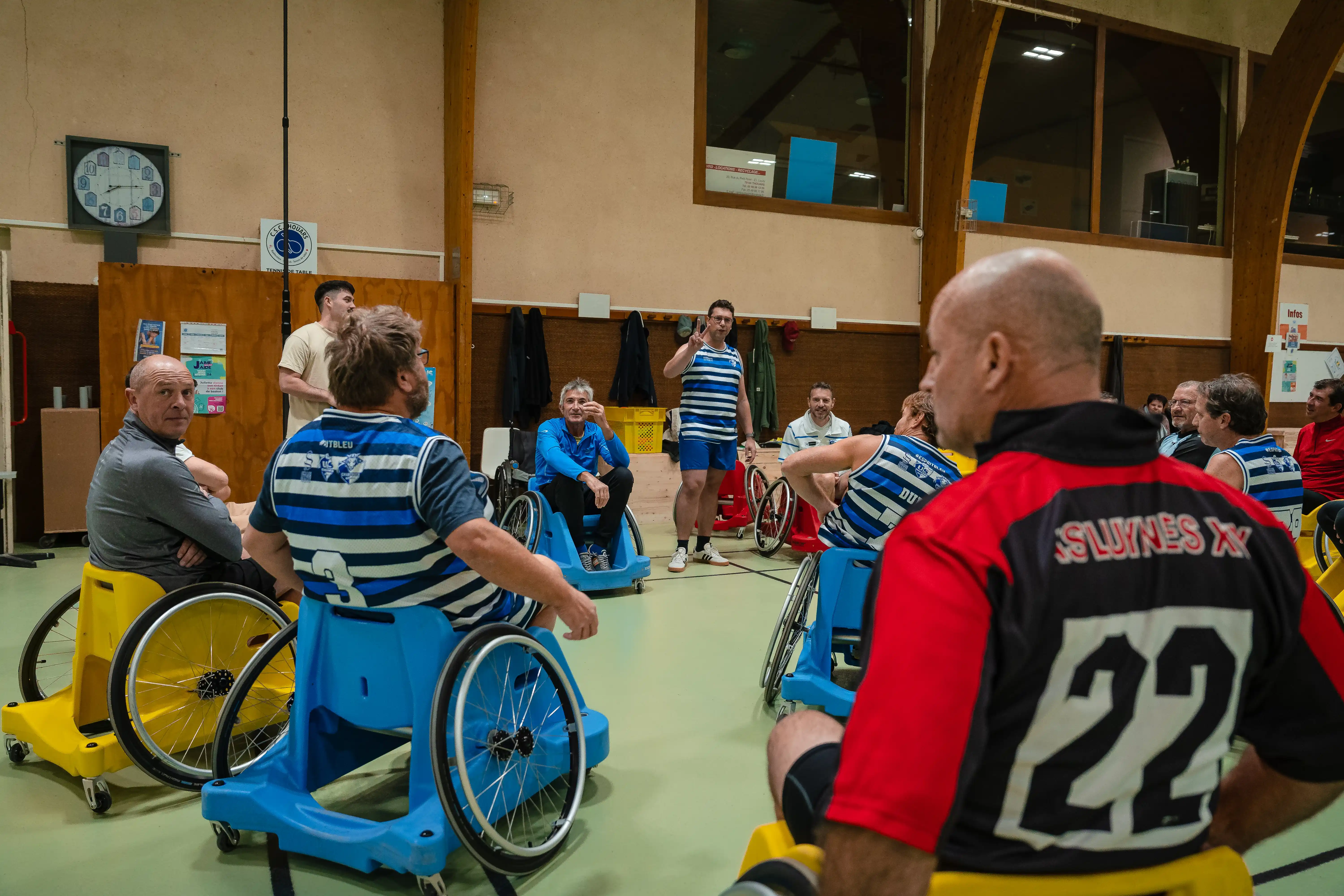 Groupe d'hommes en fauteuils roulants bleus et jaunes participant à une séance sportive en salle, certains portant des maillots rayés bleus et blancs.