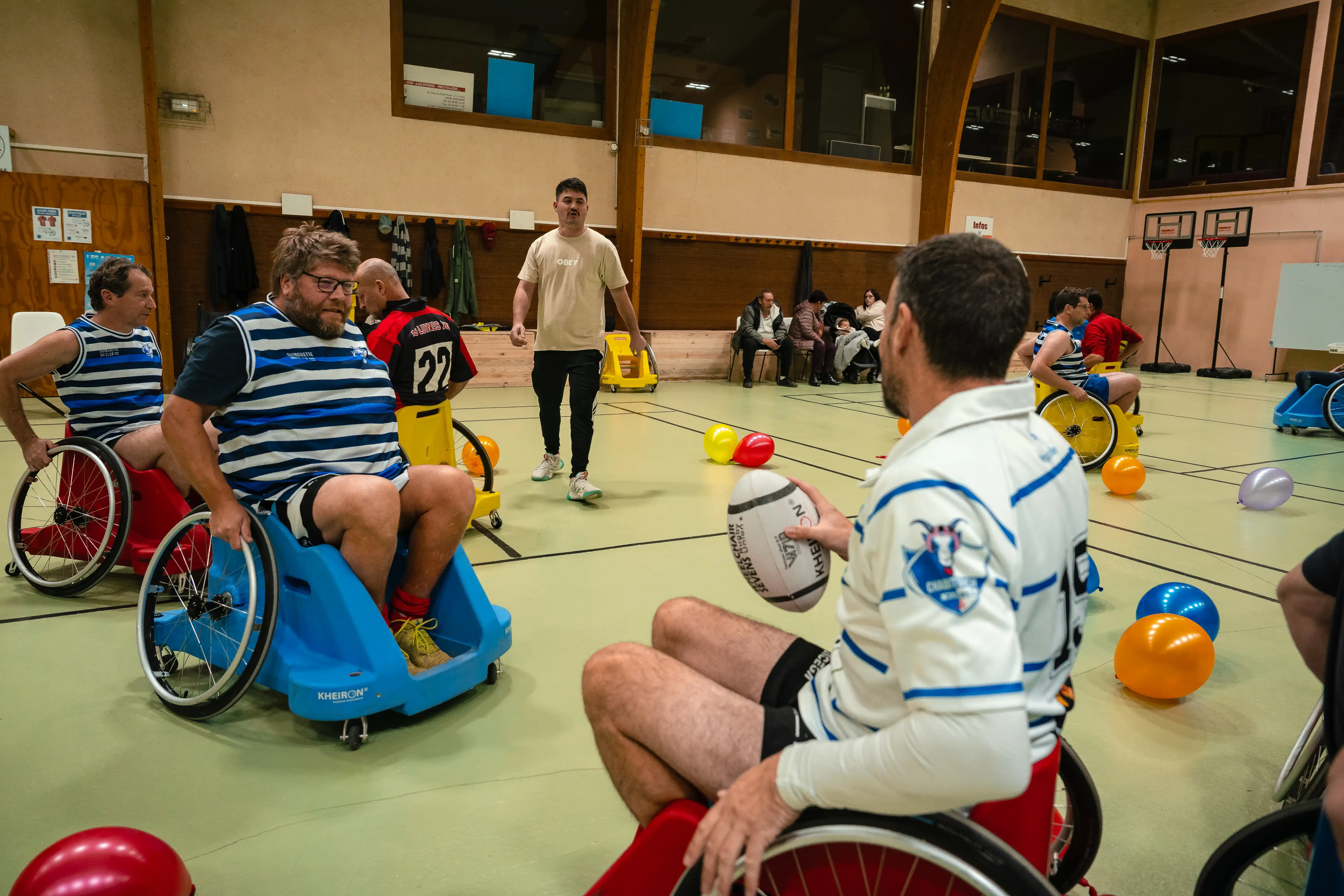Un groupe d'hommes en fauteuils roulants jouant un sport en salle, tenant un ballon ovale, entourés de ballons colorés et avec un entraîneur debout au centre.
