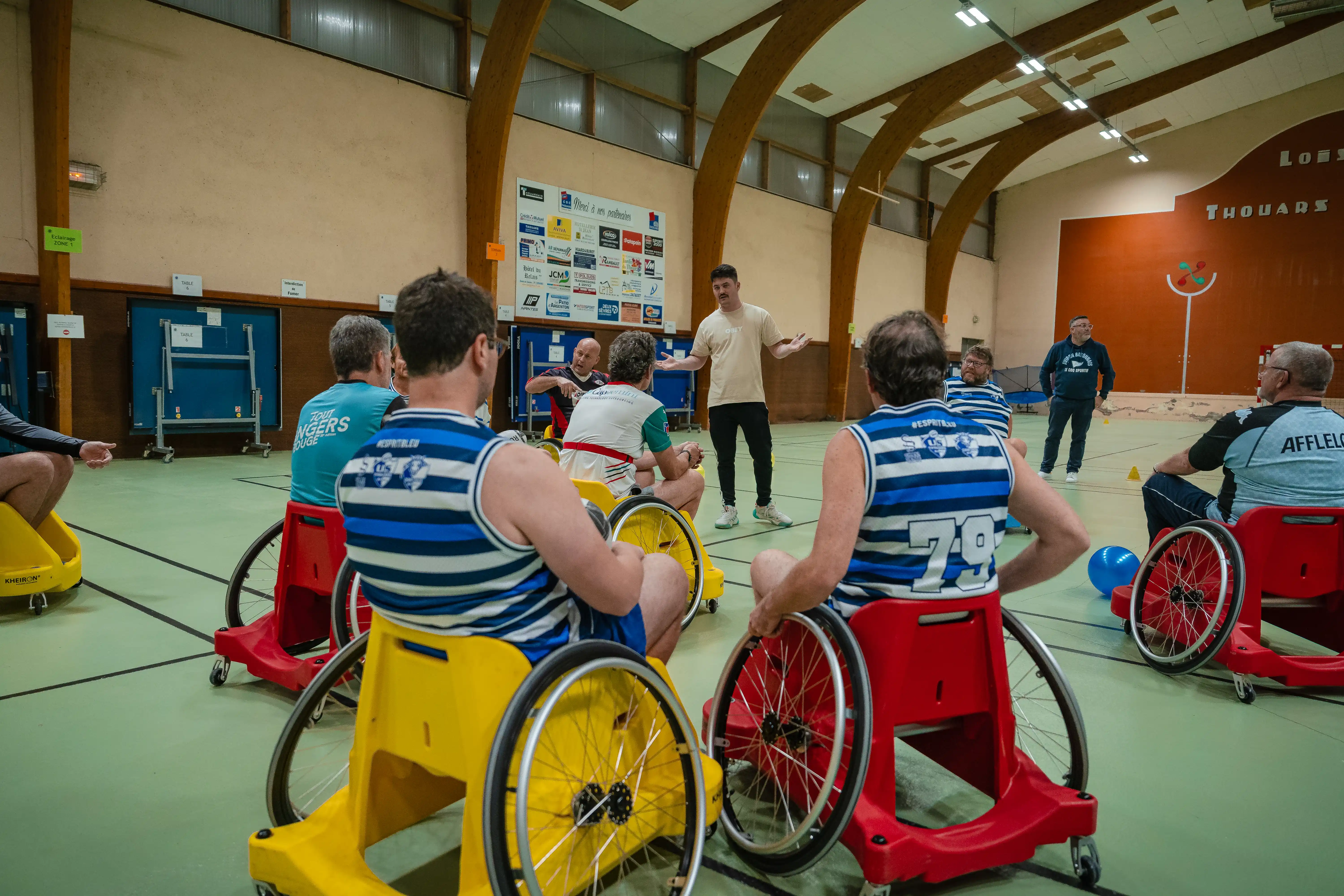 Un groupe d'hommes en fauteuils roulants sportifs écoutant un entraîneur dans une salle de sport intérieure.
