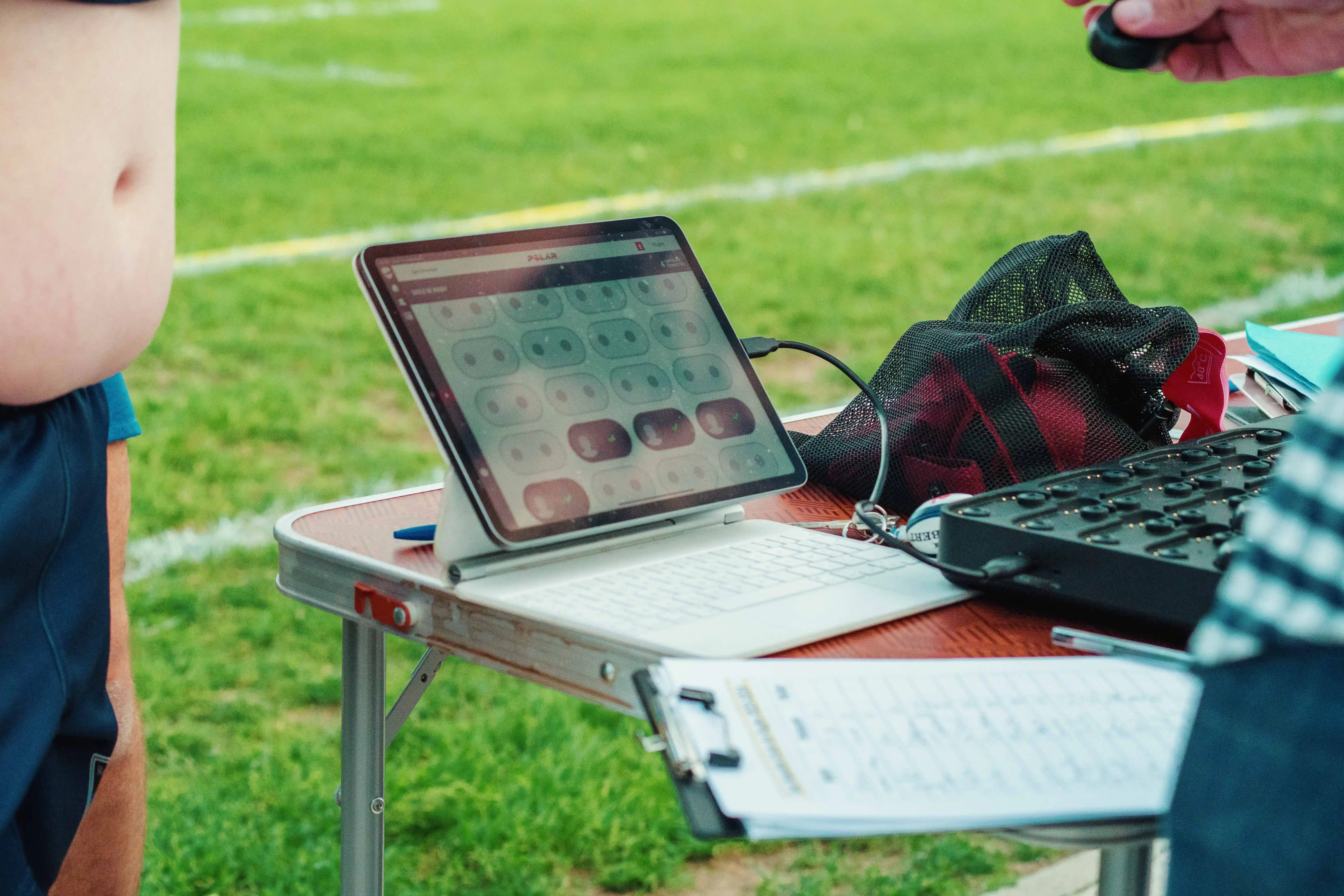 Table de terrain sportif avec une tablette affichant une application de suivi, un sac en filet rouge, et une personne tenant un bip, avec une autre personne en tenue de sport à côté.