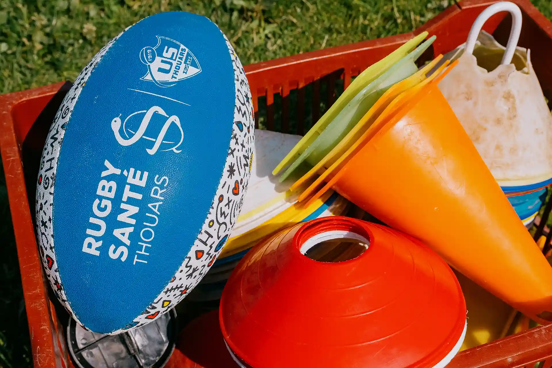 Ballon de rugby bleu marqué Rugby Sainte Thouars posé à côté de cônes d'entraînement orange et jaunes dans un panier rouge.