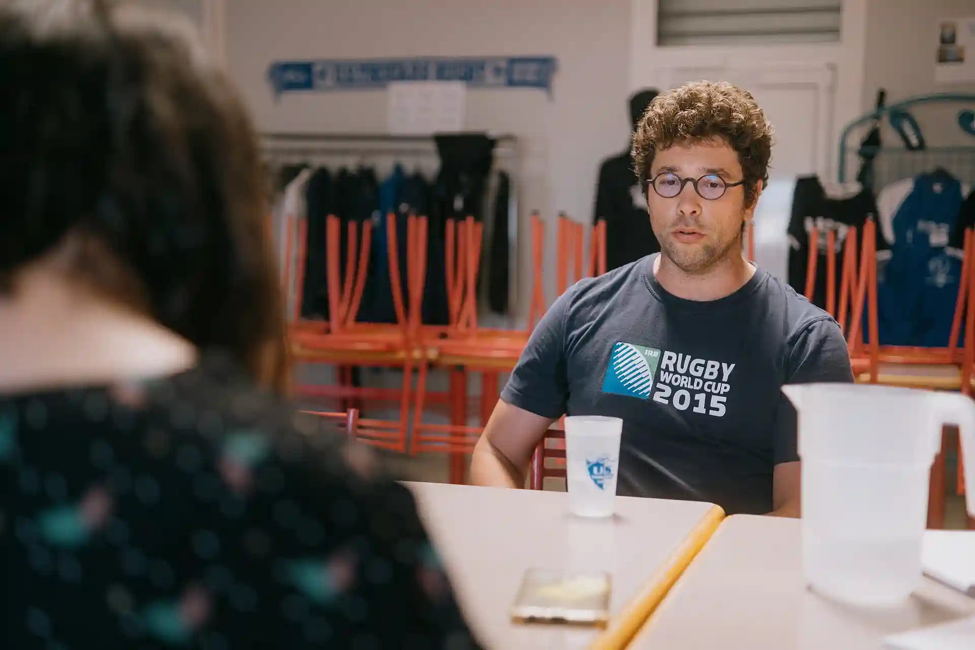Un homme portant des lunettes rondes et un t-shirt Rugby World Cup 2015, assis à une table avec un verre et une carafe d'eau, en conversation avec une personne floutée au premier plan.