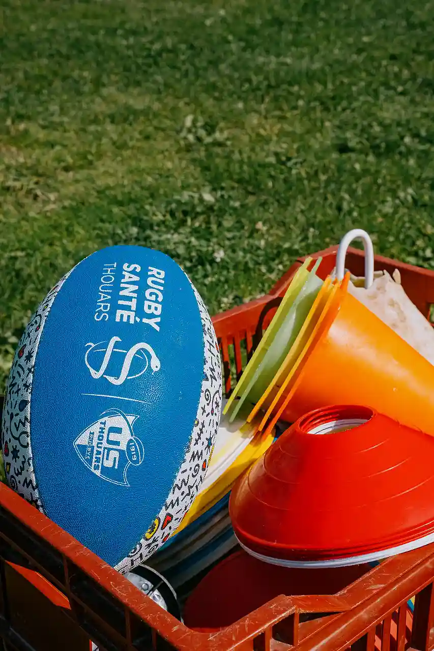 Ballon de rugby bleu avec inscription 'RUGBY SANTÉ THOUARS' dans une caisse avec cônes et plots d'entraînement sur herbe.
