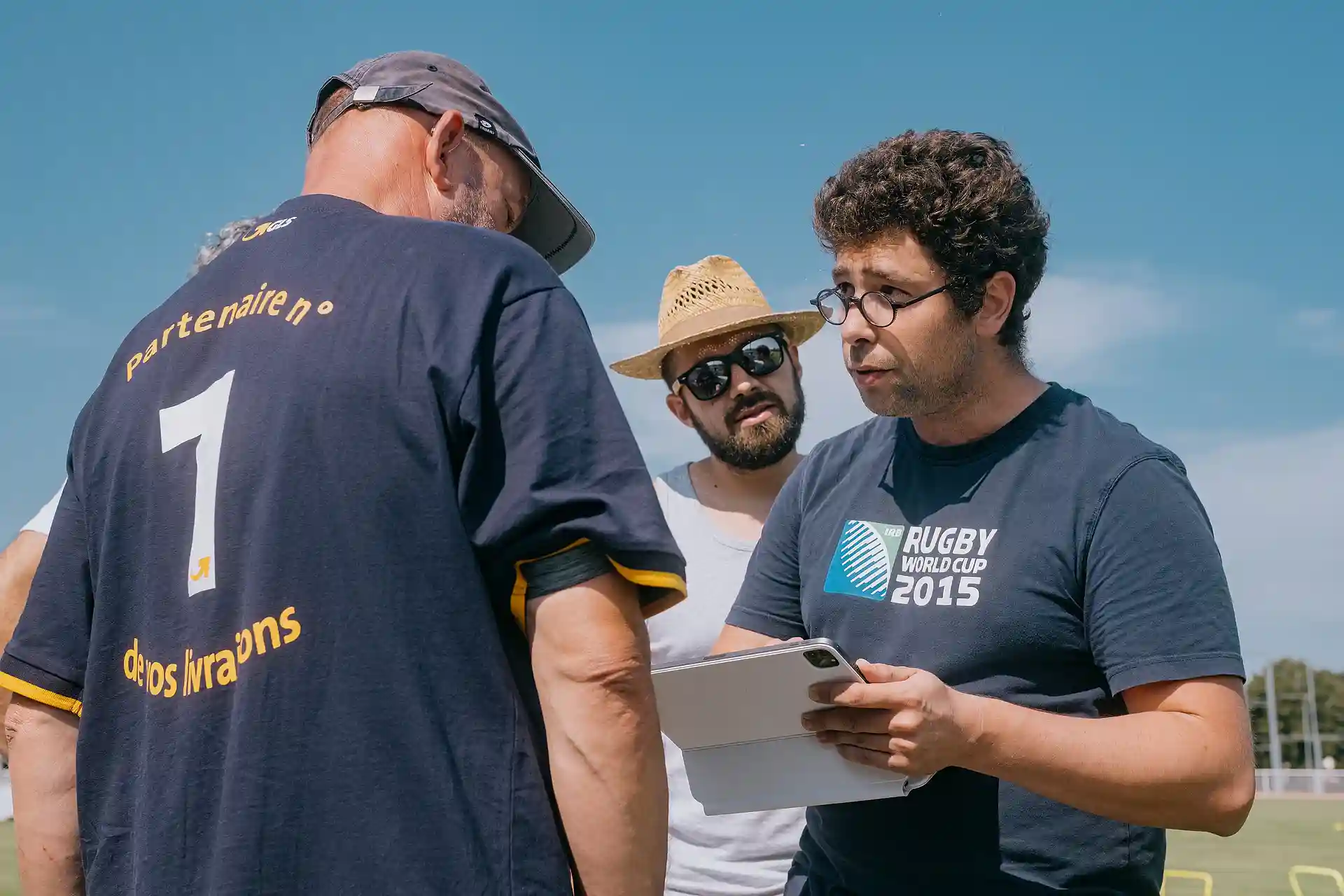 Trois hommes discutant à l'extérieur, l'un portant un t-shirt Rugby World Cup 2015 et tenant une tablette.