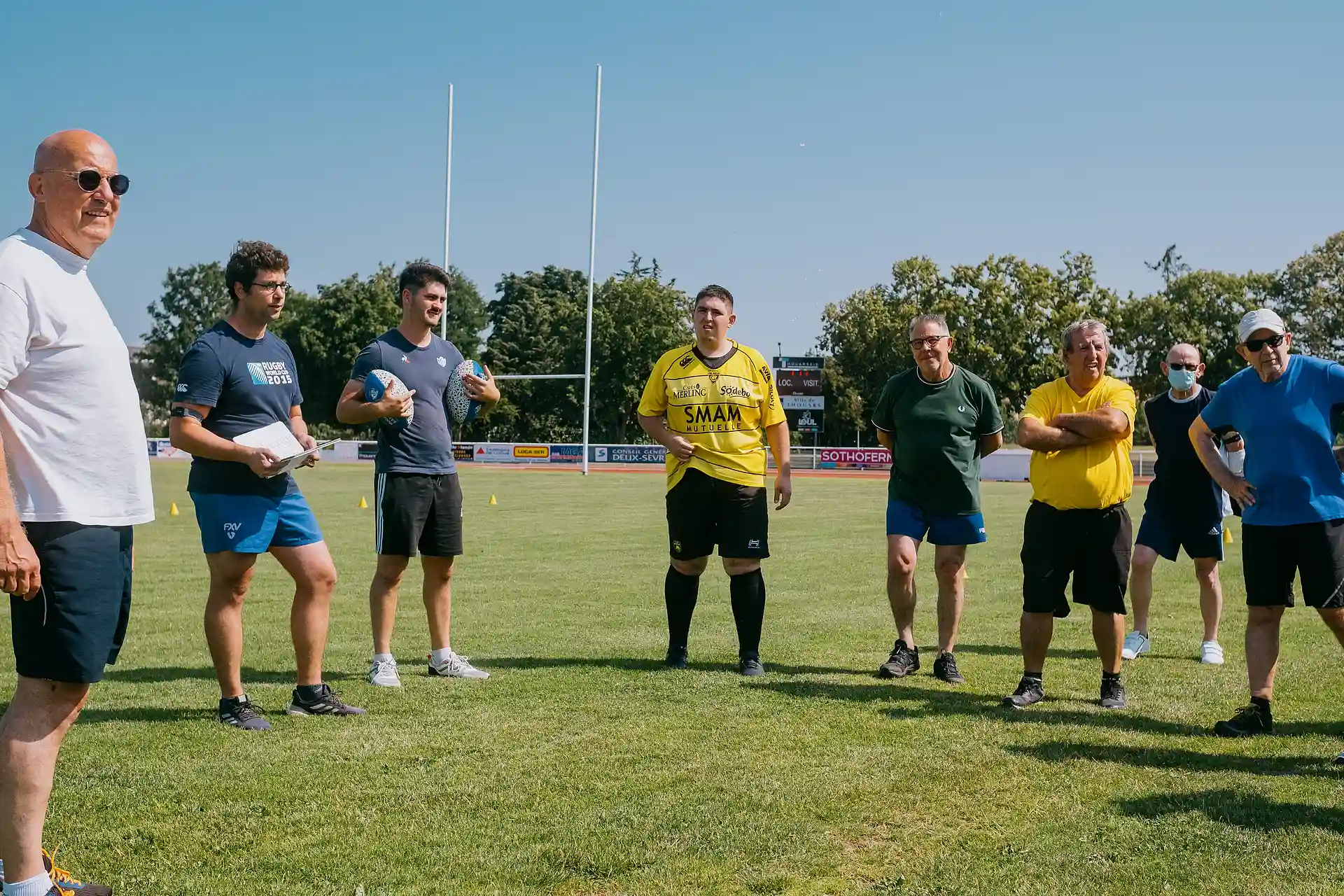 Un groupe d'hommes de différents âges debout sur un terrain de rugby sous un ciel bleu clair.