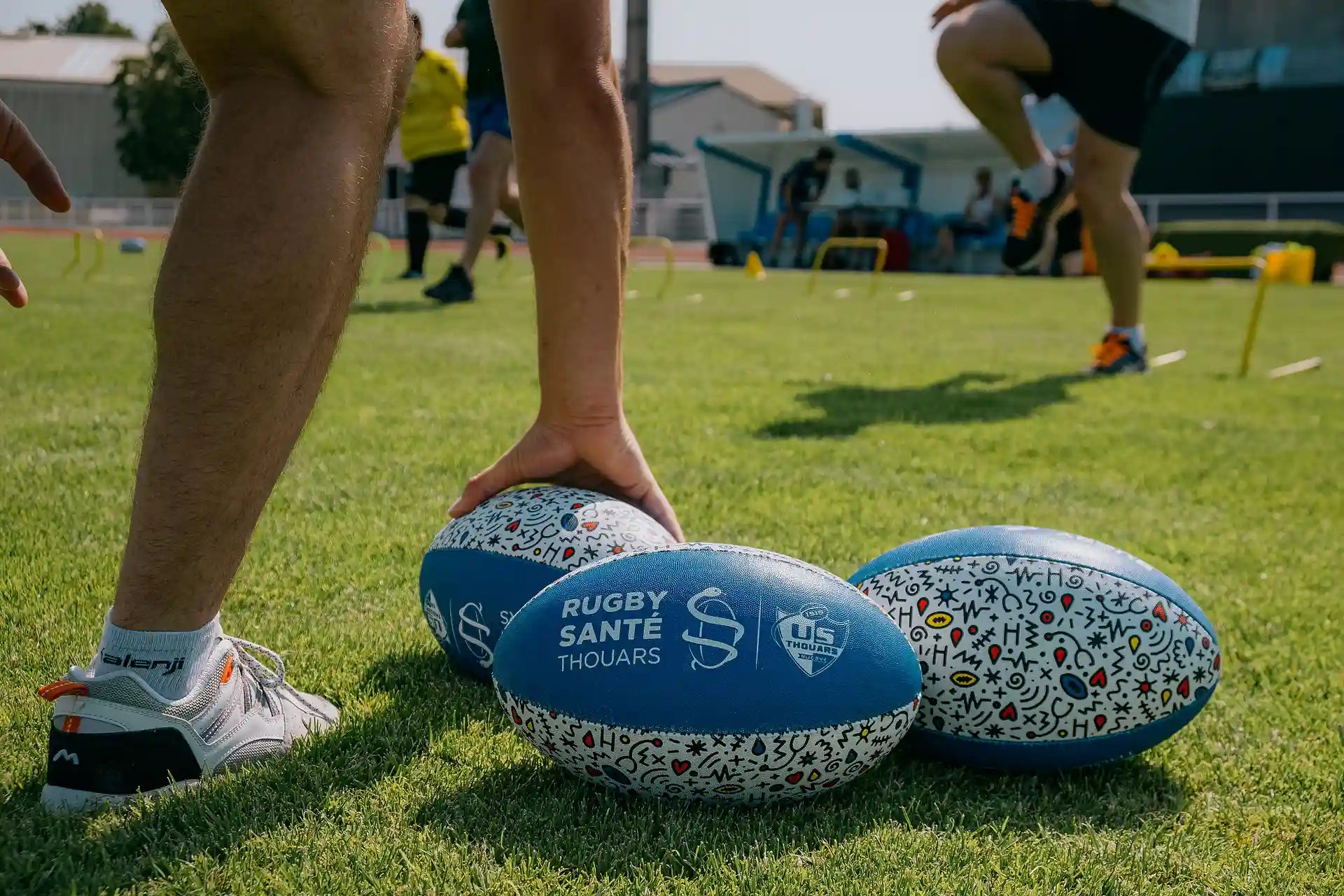Un joueur tenant un ballon de rugby marqué 'Rugby Santé Thouars' sur un terrain d'entraînement avec plusieurs ballons et des joueurs en arrière-plan.