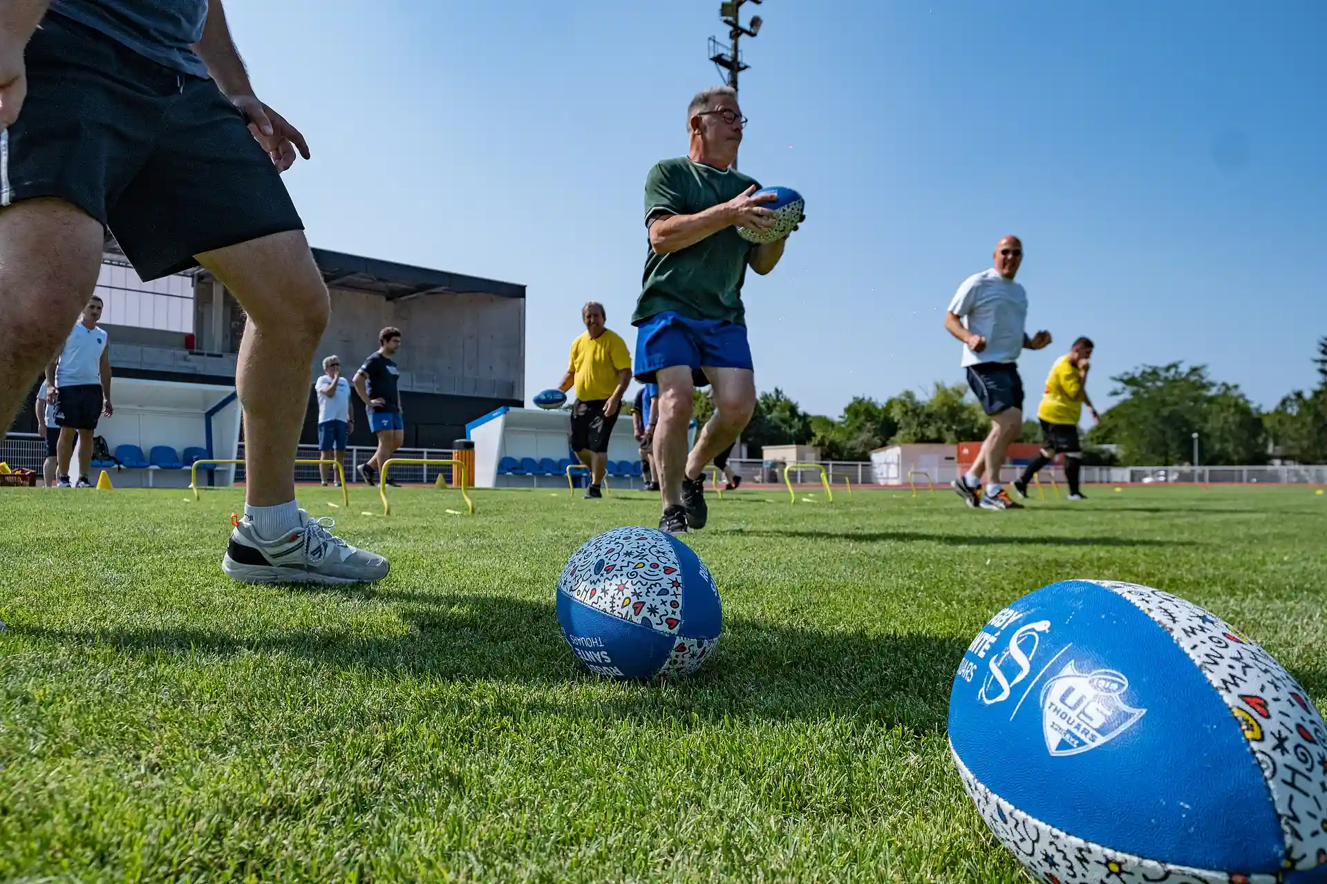 Un groupe d'adultes s'entraînant au rugby en extérieur sur un terrain herbeux, avec plusieurs ballons de rugby colorés au premier plan.