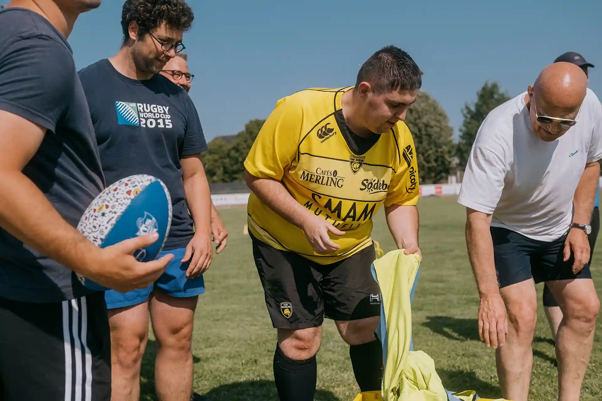 Quatre hommes sur un terrain de rugby, dont un avec un ballon de rugby en main et un autre portant un maillot jaune et noir.