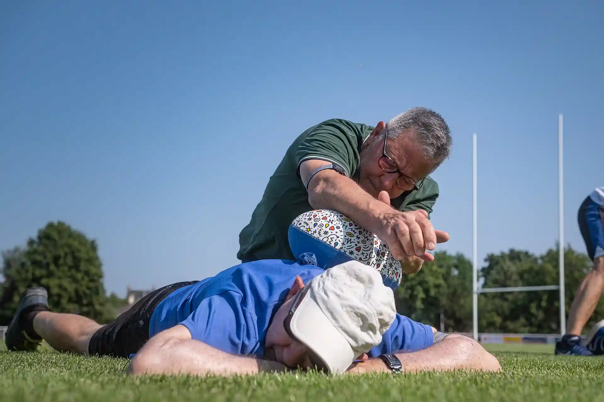 Un homme en polo vert avec des lunettes tient un ballon de rugby sur le dos d'un homme allongé portant une casquette blanche sur un terrain de rugby ensoleillé.