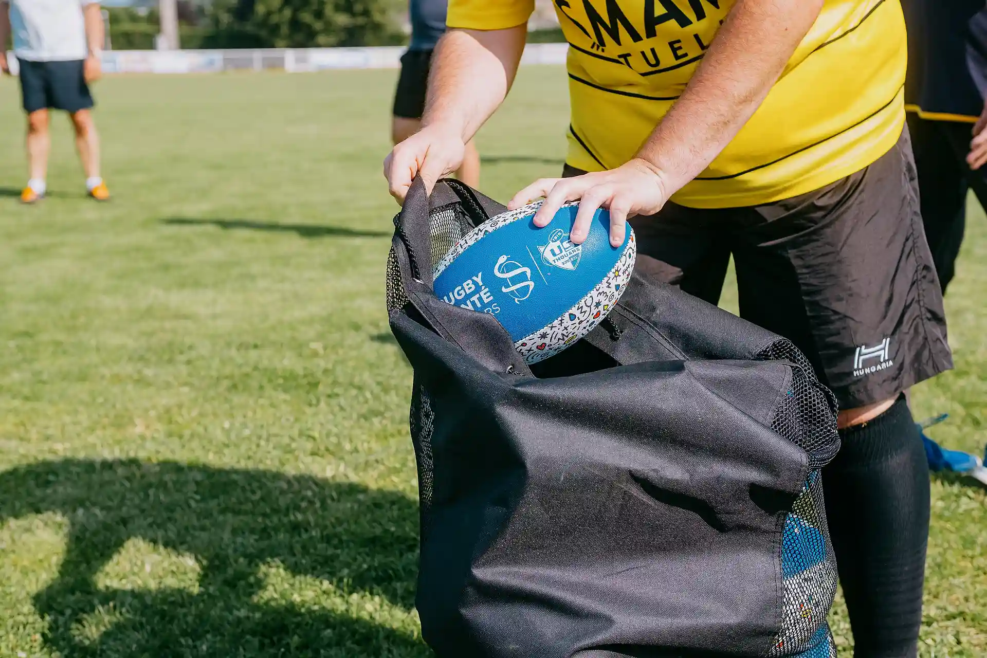 Une personne en maillot jaune range un ballon de rugby bleu dans un sac noir sur un terrain de sport herbeux.
