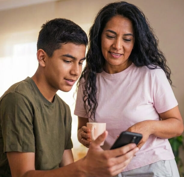 A teenage boy shows something on his smartphone to a smiling woman holding a coffee mug.