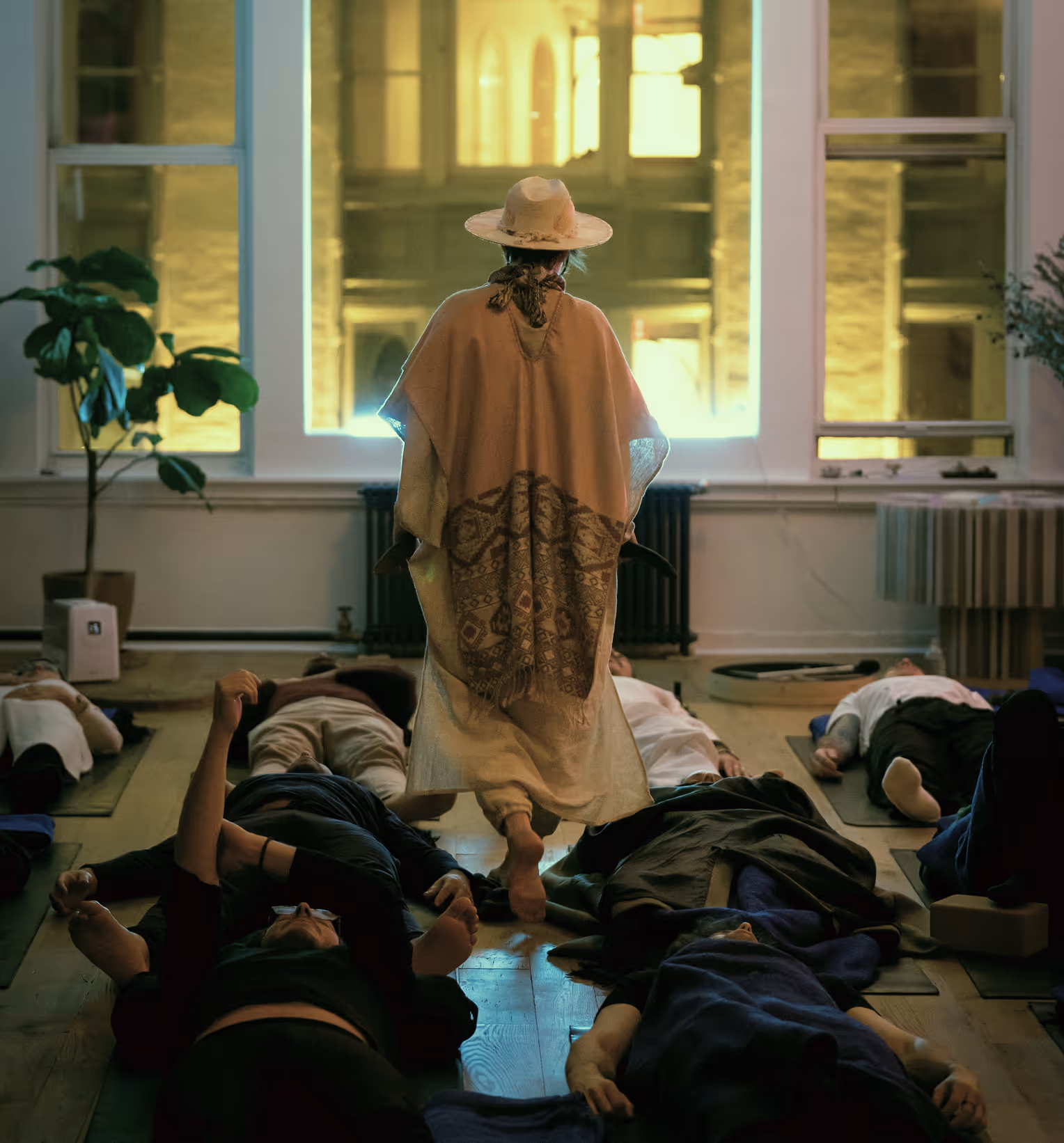 Instructor wearing a hat and poncho walking barefoot among people lying on yoga mats in a dimly lit room with large windows.