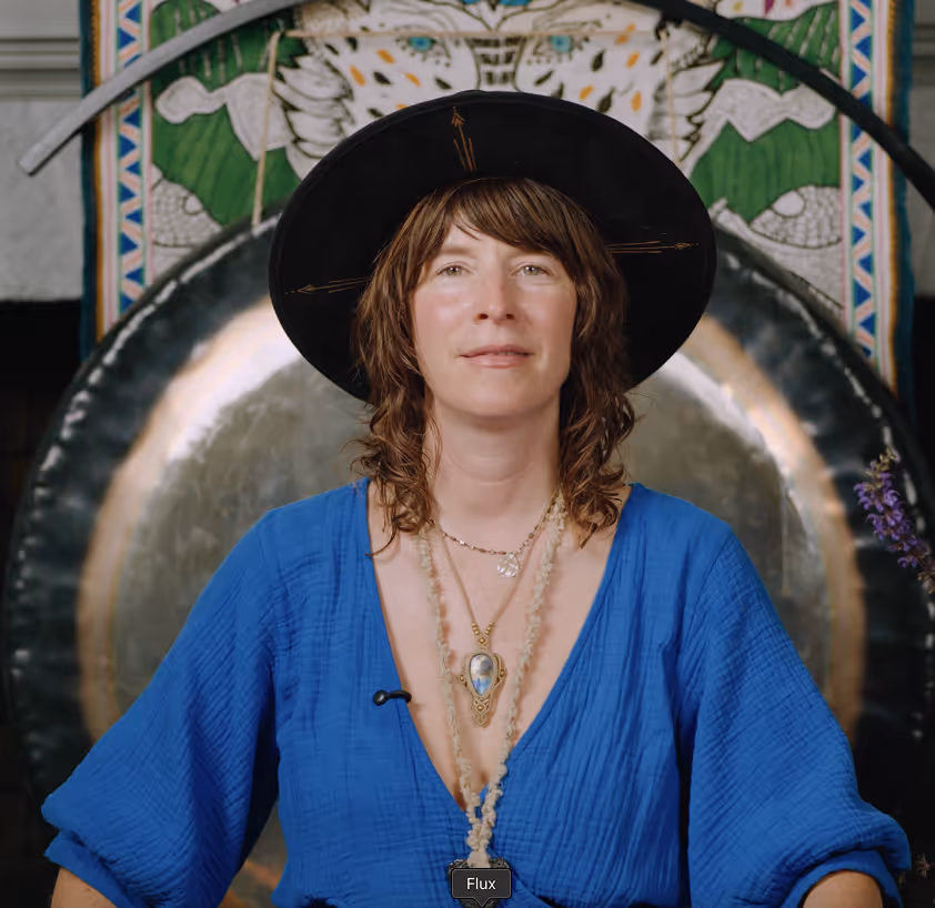 Woman with shoulder-length brown hair wearing a black wide-brimmed hat and blue top, sitting in front of a large circular gong.