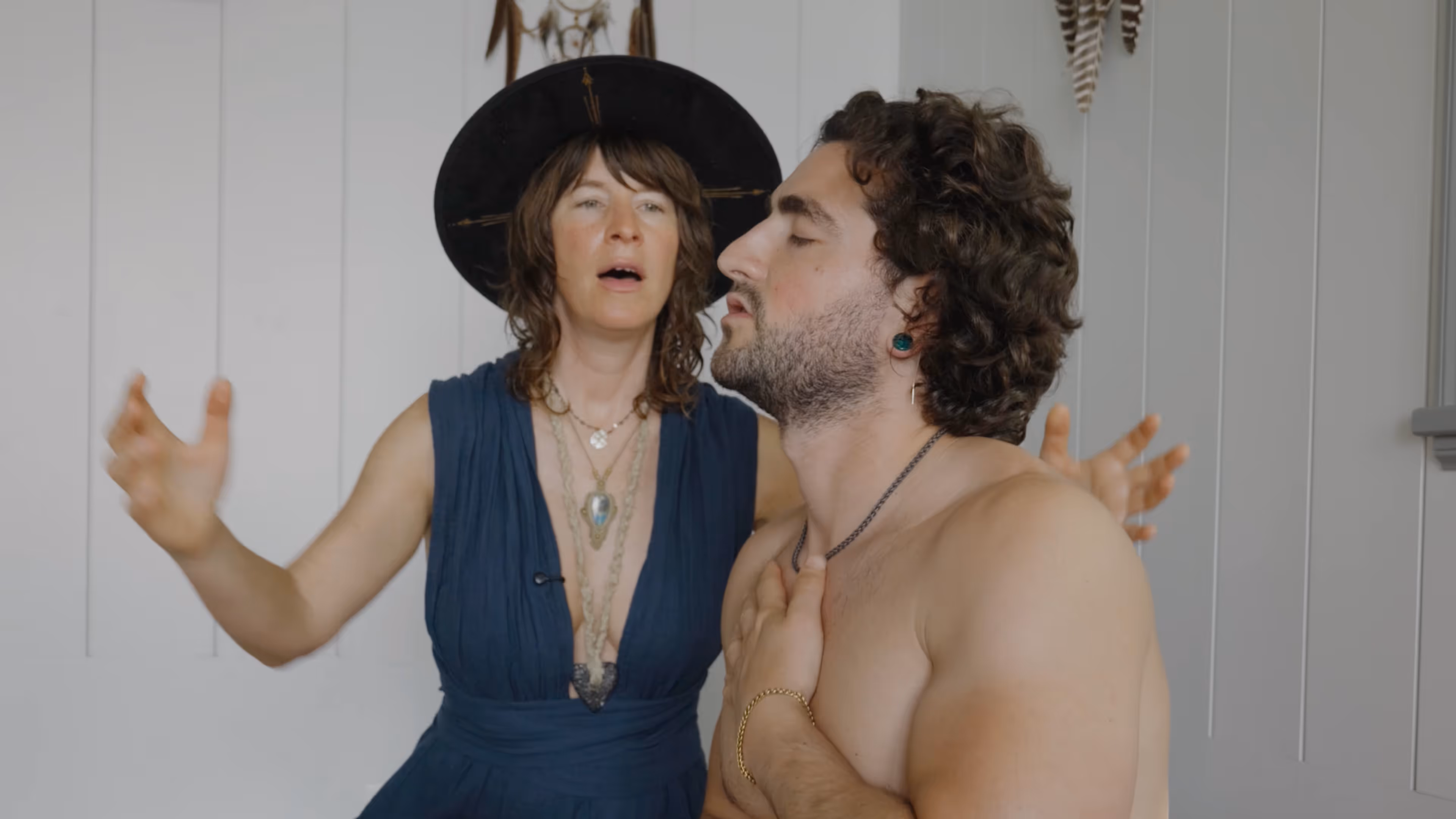 A woman wearing a black hat and blue dress gestures with her hands behind a shirtless man with curly hair and closed eyes holding his chest.