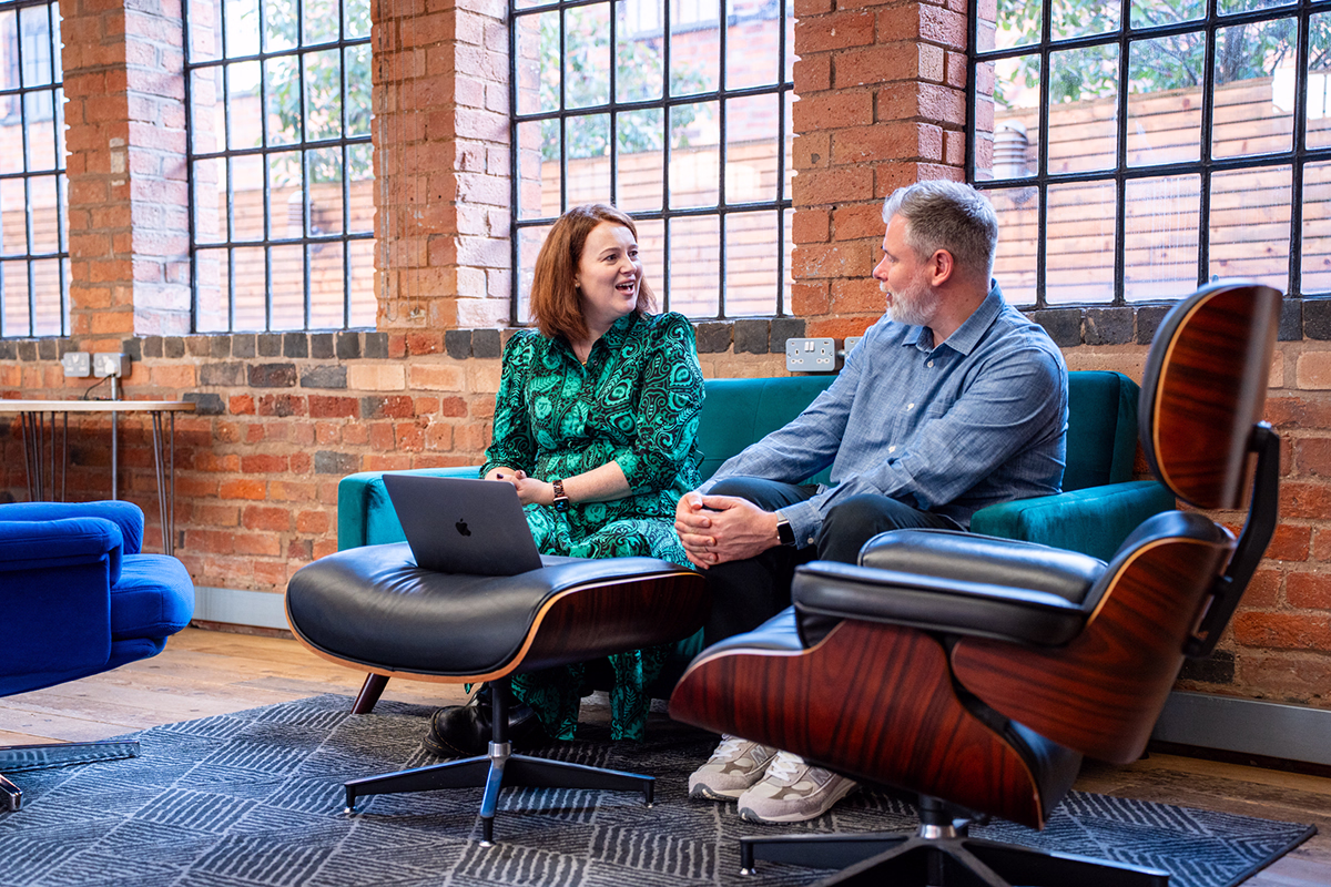 Two people sitting on a green couch in a modern brick-walled office, engaged in conversation with a laptop on a leather ottoman in front of them.