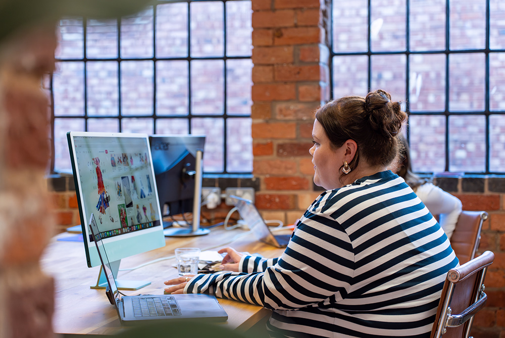 Woman in a striped shirt working on a desktop computer in a brick-walled office with large windows.