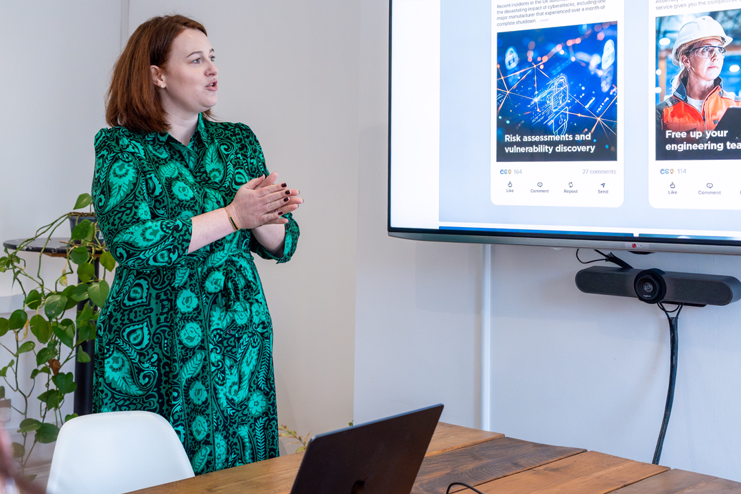 Woman in a green patterned dress giving a presentation in a meeting room with a large screen displaying cybersecurity and engineering content.