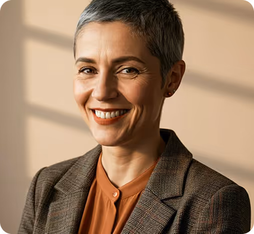 Smiling middle-aged woman with short gray hair wearing a brown blazer and rust-colored blouse against a beige background.