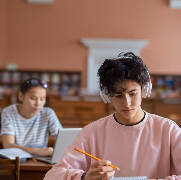 Young man wearing headphones and a pink sweatshirt writing with a pencil in a notebook in a library setting, with another person working on a laptop in the background.