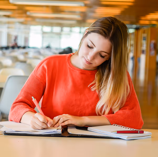 Young woman in red sweater writing in a notebook at a library or study area.