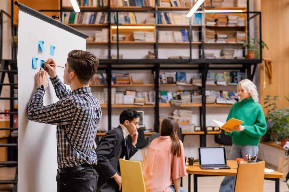 Young man placing blue sticky notes on a whiteboard while three colleagues work and discuss in a modern office with shelves full of books.