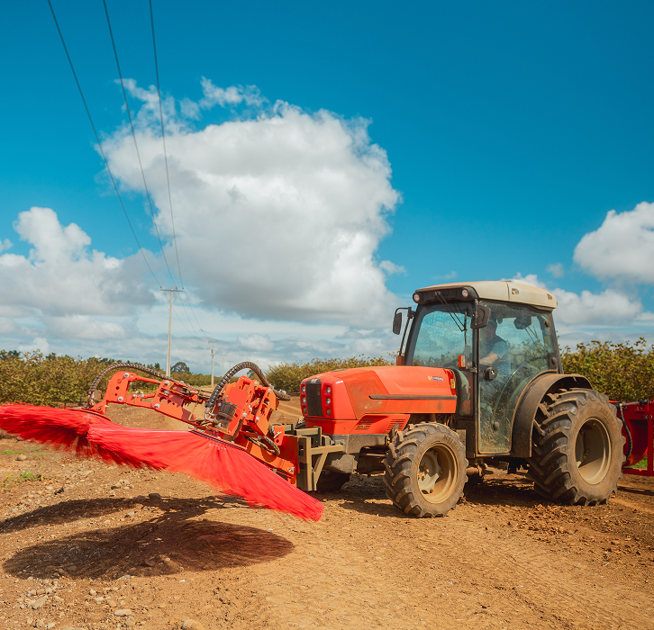 Orange tractor with a large rotating red brush attachment on a dirt path under a blue sky with white clouds.