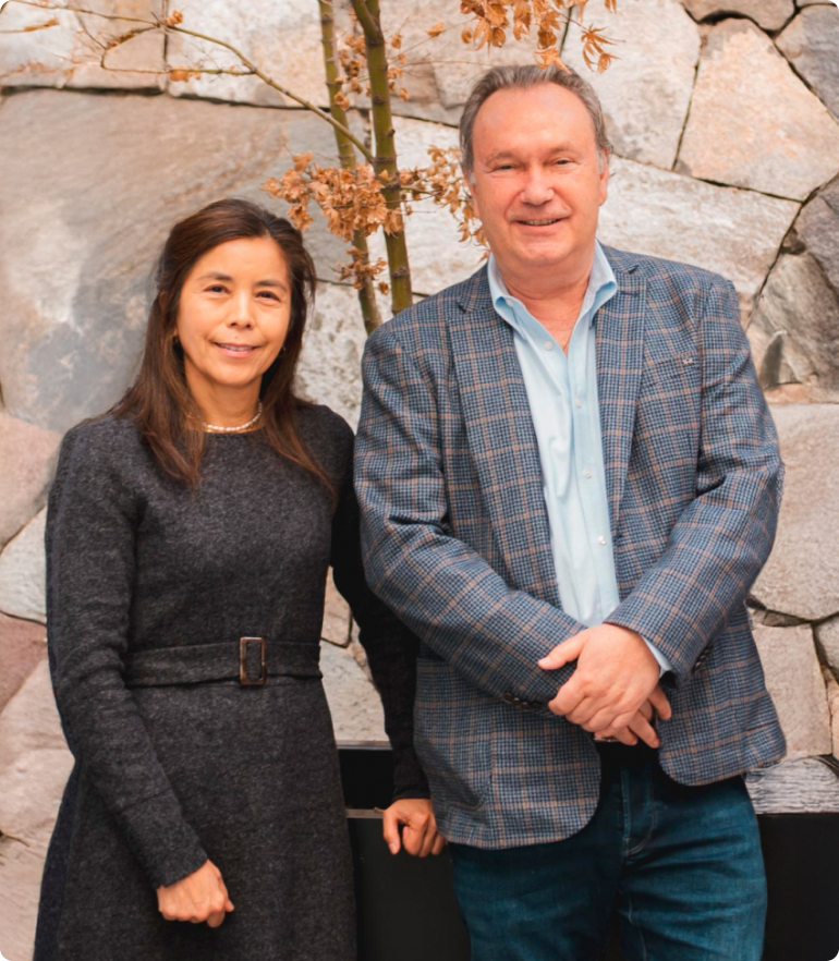 Smiling man in a plaid blazer and woman in a dark dress standing side by side in front of a stone wall with a small tree.