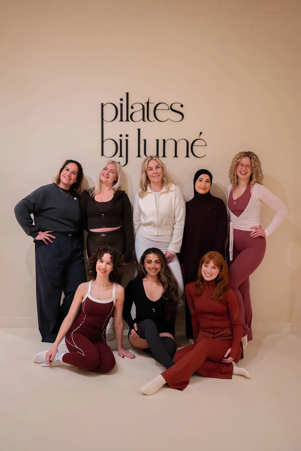 Een groep van zeven diverse vrouwen lacht en poseert in sportkleding voor een beige muur met de tekst 'pilates bij lume'.
