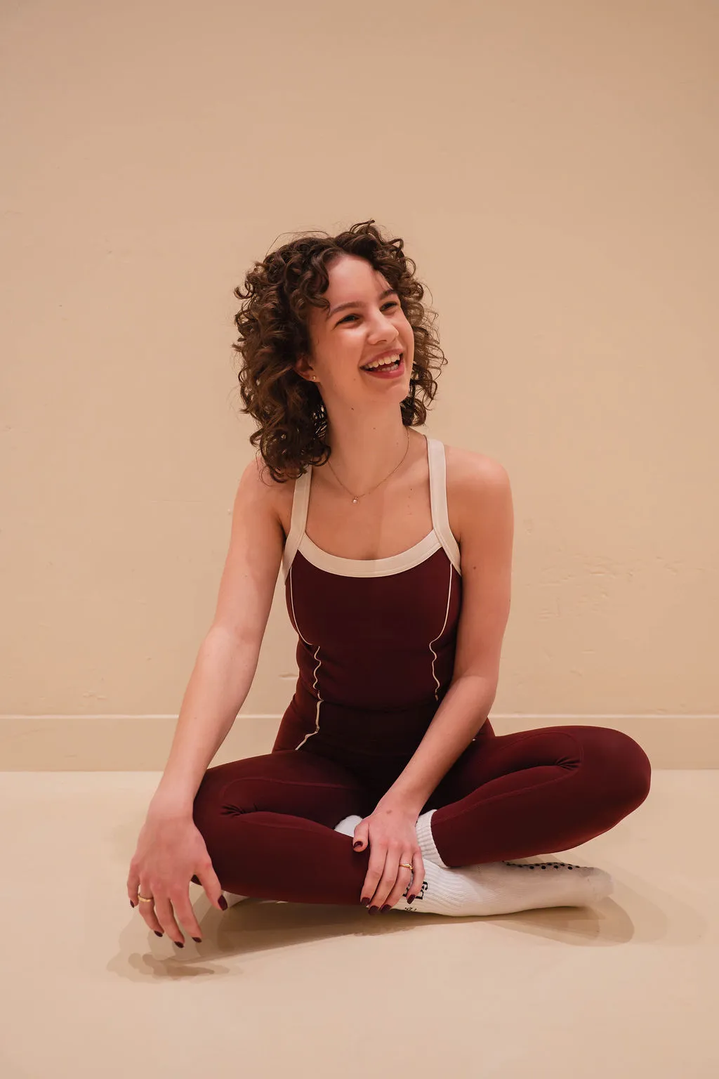 Lachende vrouw met krullend haar in bordeauxrode sportkleding zit met gekruiste benen op een lichte vloer tegen een beige muur.