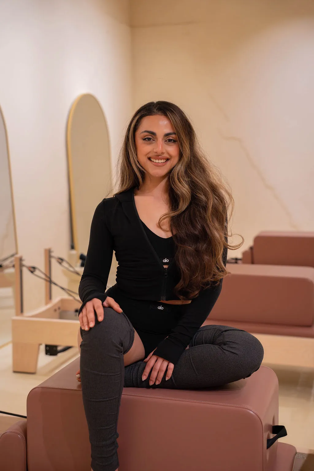 Lachende vrouw met lang golvend haar zit met gekruiste benen op een mauvekleurig pilatesapparaat in een lichte studio.