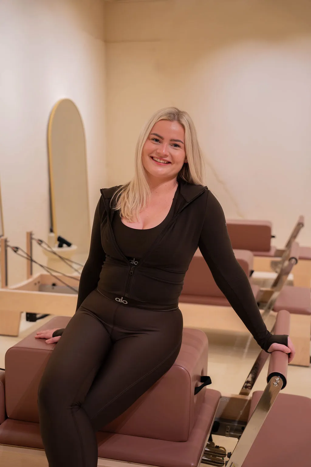 Lachende vrouw in zwarte sportkleding zit op een Pilates reformer in de studio.