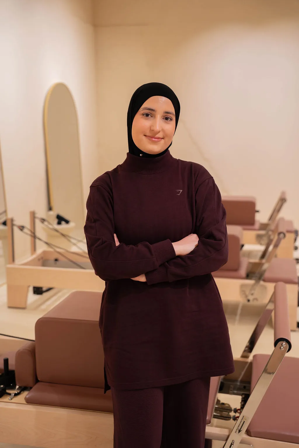 Een lachende vrouw met een zwarte hijab en een donkerrode outfit staat met haar armen over elkaar in een pilatesstudio.